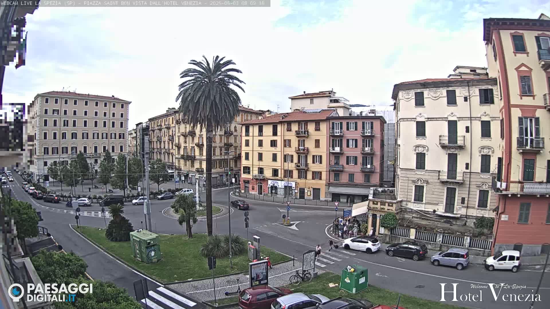 A mostly sunny, outdoor view of a European city square with several multi-story buildings, cars, and a palm tree in the center of a roundabout.