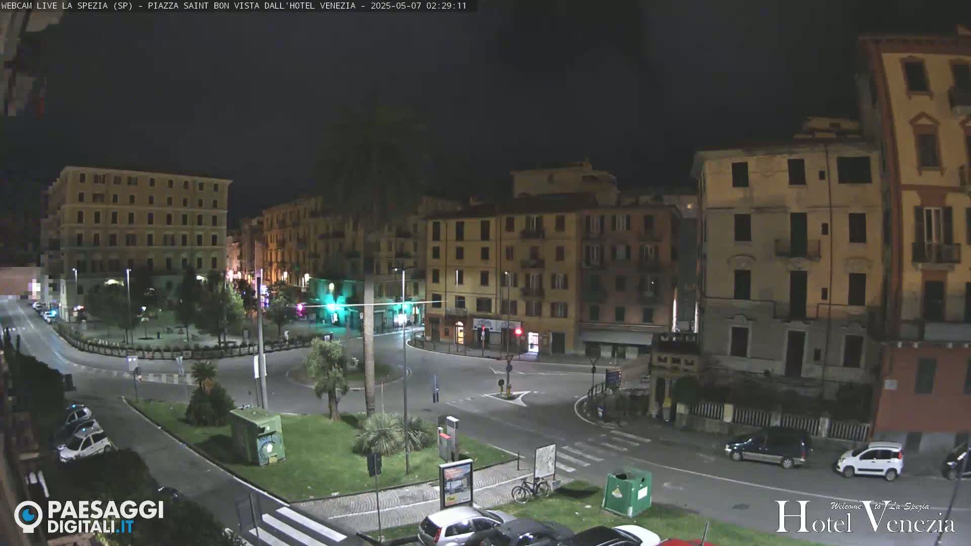 A nighttime view of a mostly empty town square surrounded by multi-story buildings, with a few cars parked and driving.