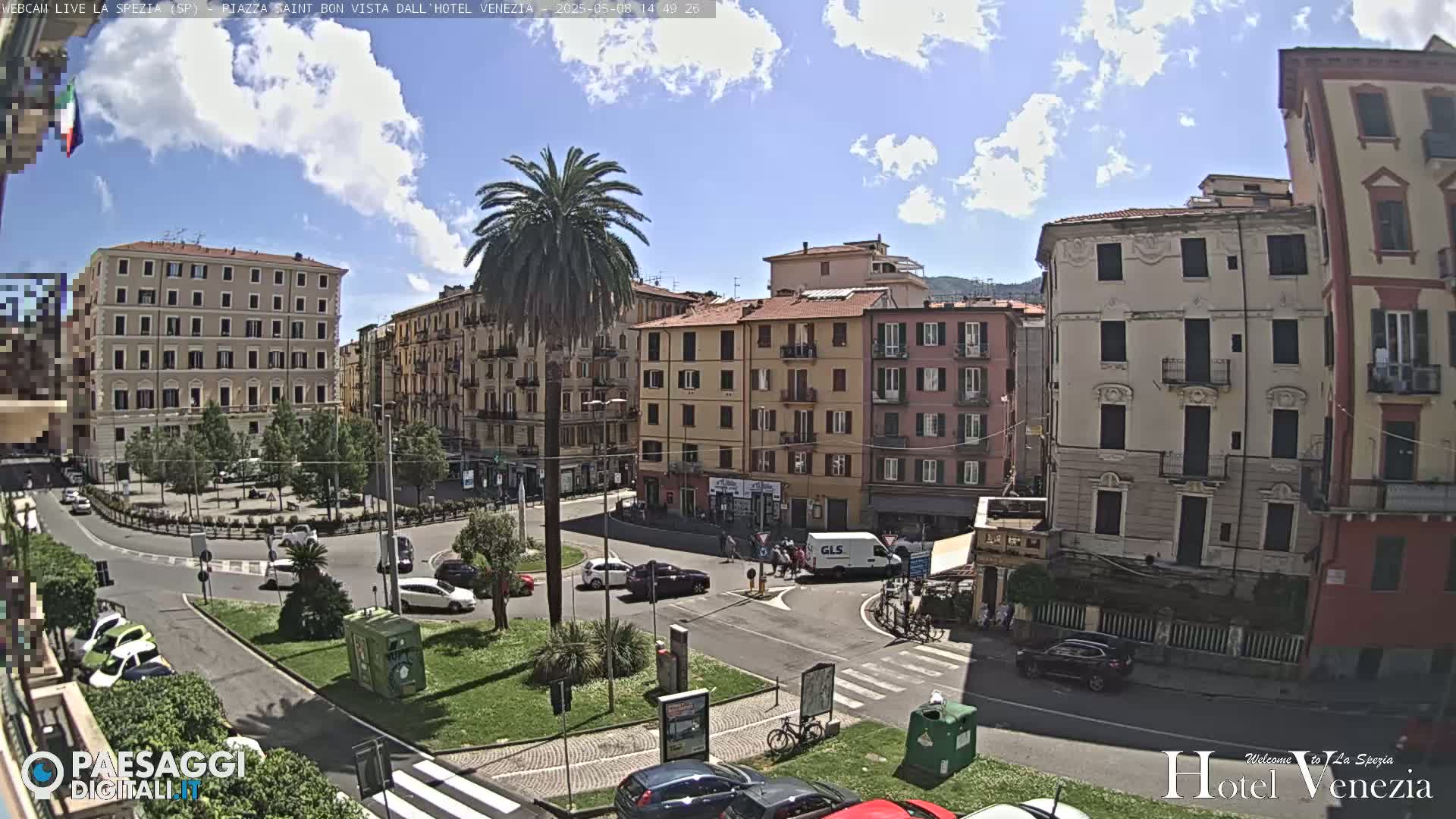 A sunny day in a European city square shows several multi-story buildings surrounding a roundabout with cars and a palm tree in the center.