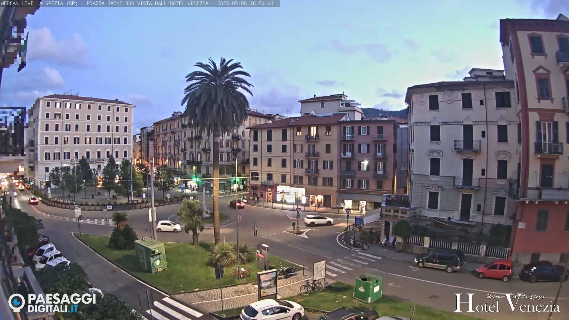 A twilight view of a city square surrounded by multi-story buildings, with a few cars and a palm tree in the center.