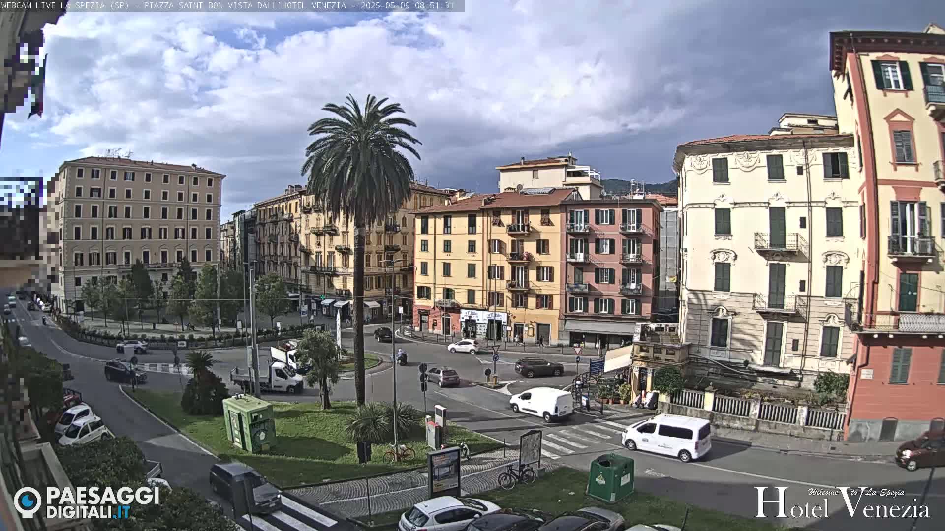 A partly cloudy day reveals a European city square with several multi-story buildings, a central palm tree, and light traffic.