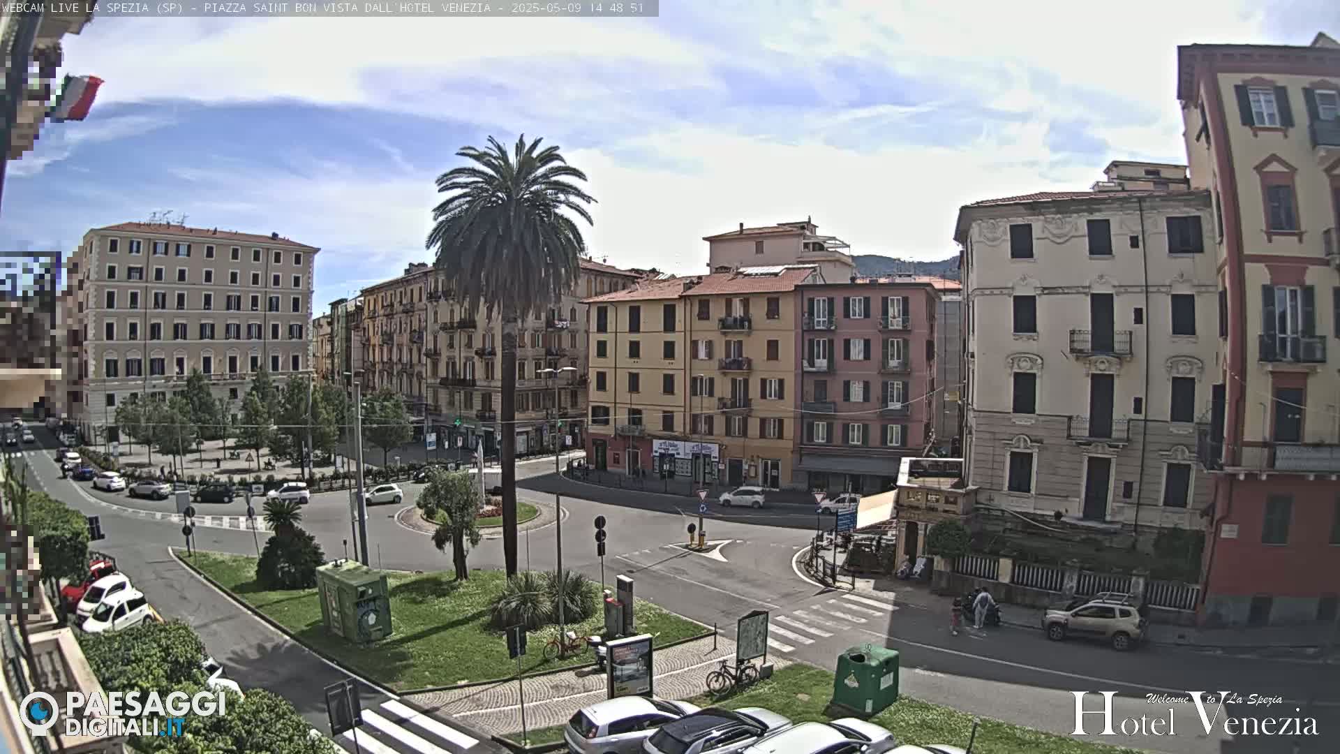 A sunny, partly cloudy day in a European town square shows several multi-story buildings surrounding a circular traffic intersection, a central palm tree, and parked cars.