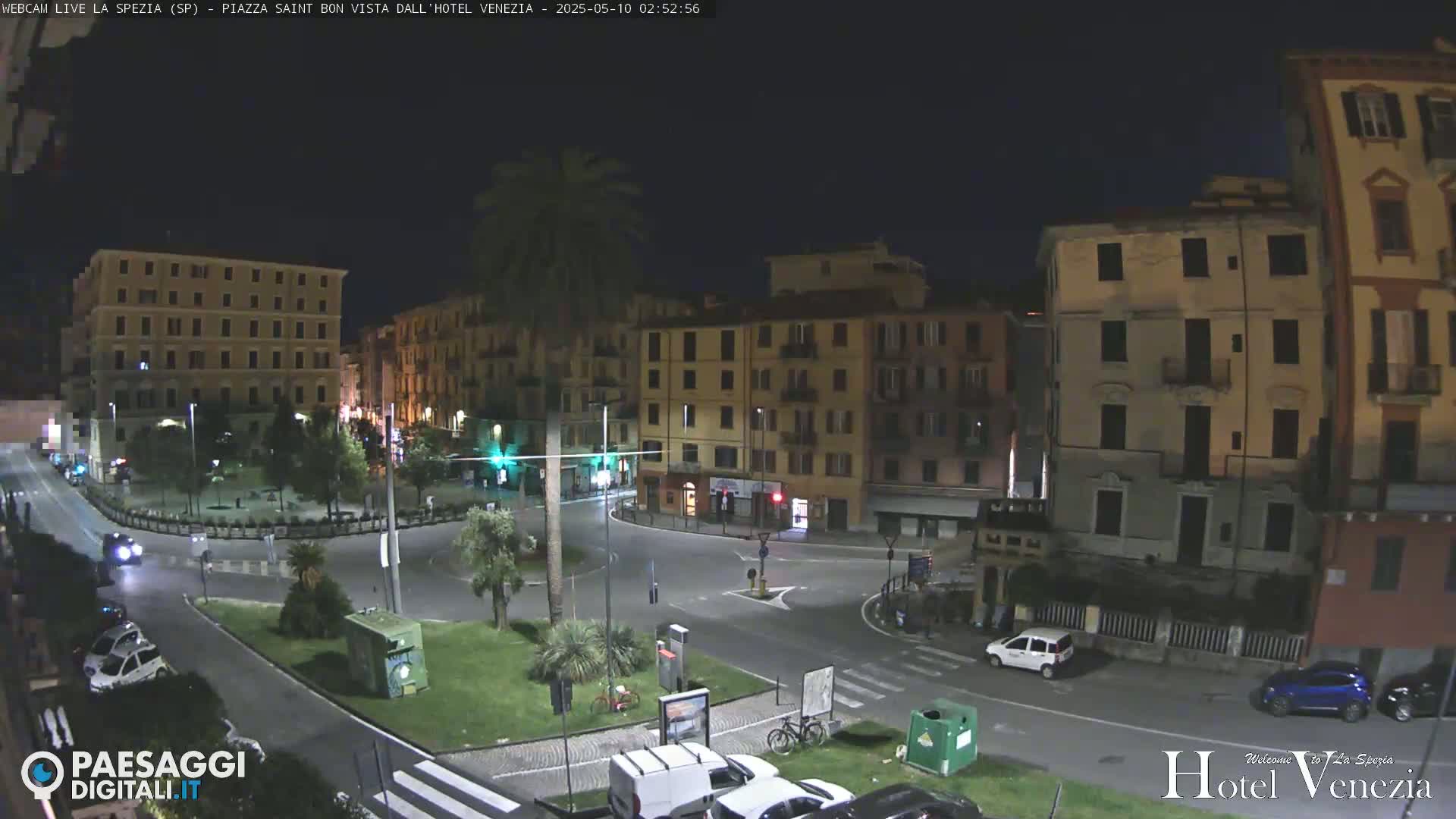 A nighttime, high-angle view of a quiet town square, surrounded by multi-story buildings, with a few cars and a bicycle parked nearby.