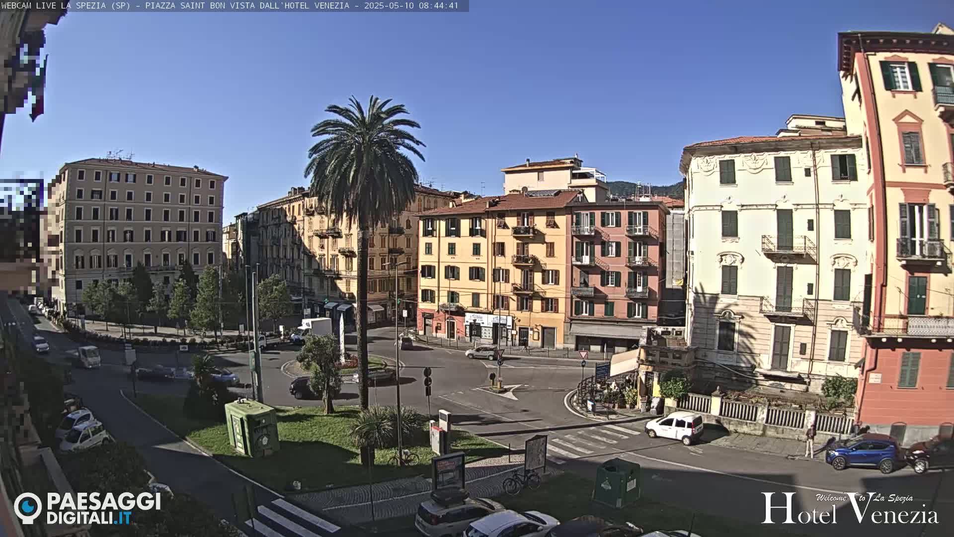 A sunny cityscape view shows a traffic circle surrounded by multi-story buildings, palm trees, and cars.