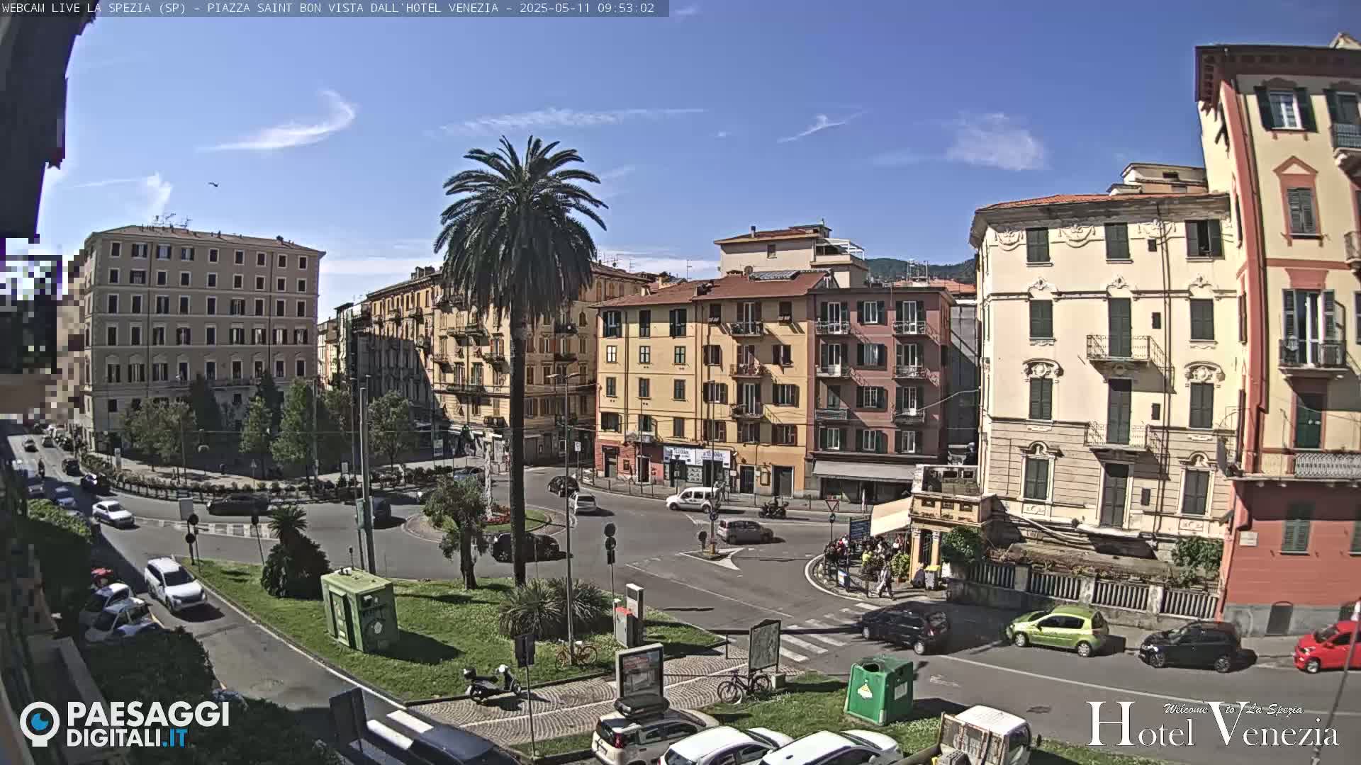 A sunny day in a European city square, showing multi-story buildings, palm trees, and light traffic.