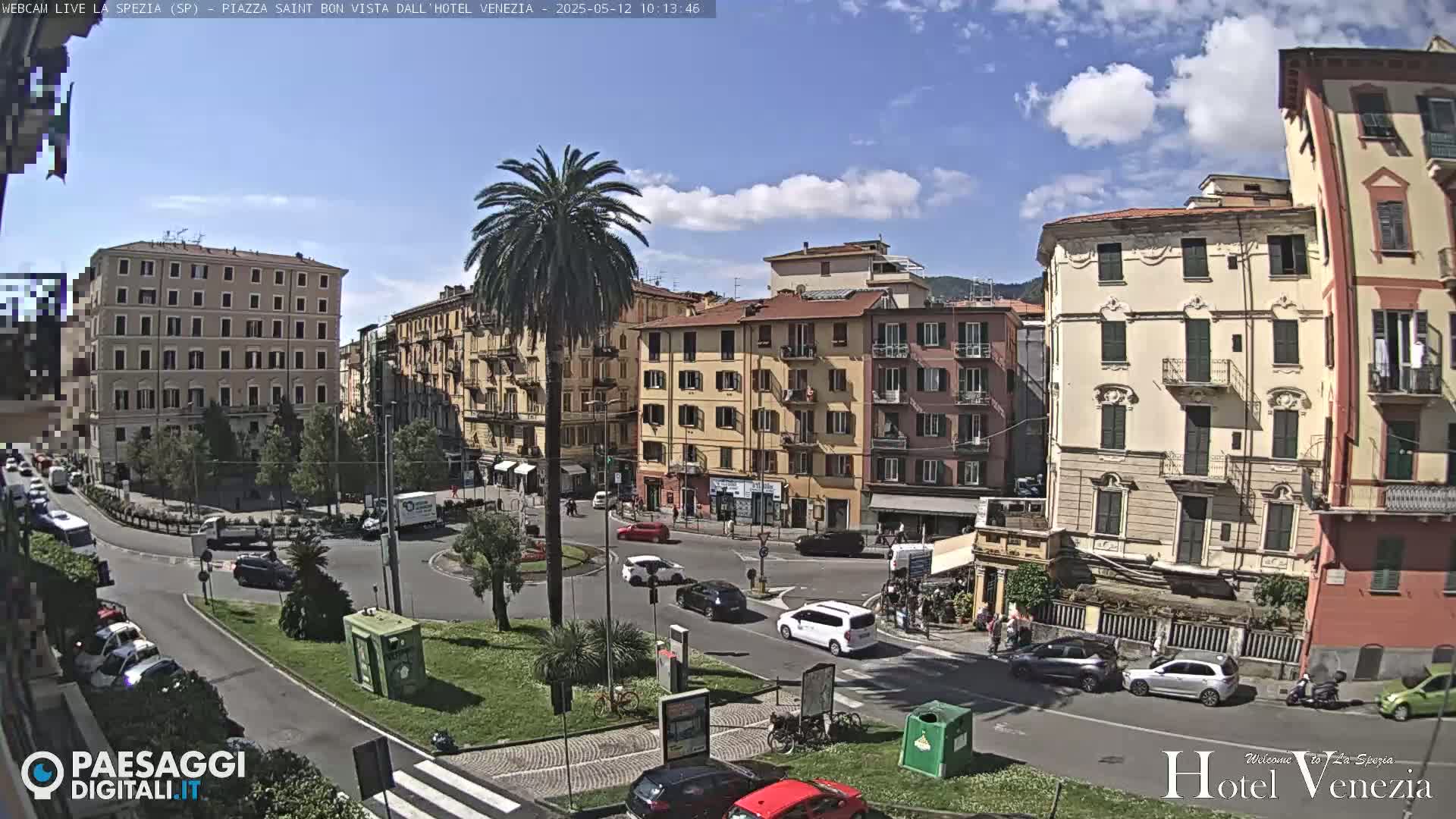 A sunny day in a European town square, where several multi-story buildings surround a roundabout with cars and pedestrians, and a tall palm tree stands in the center.