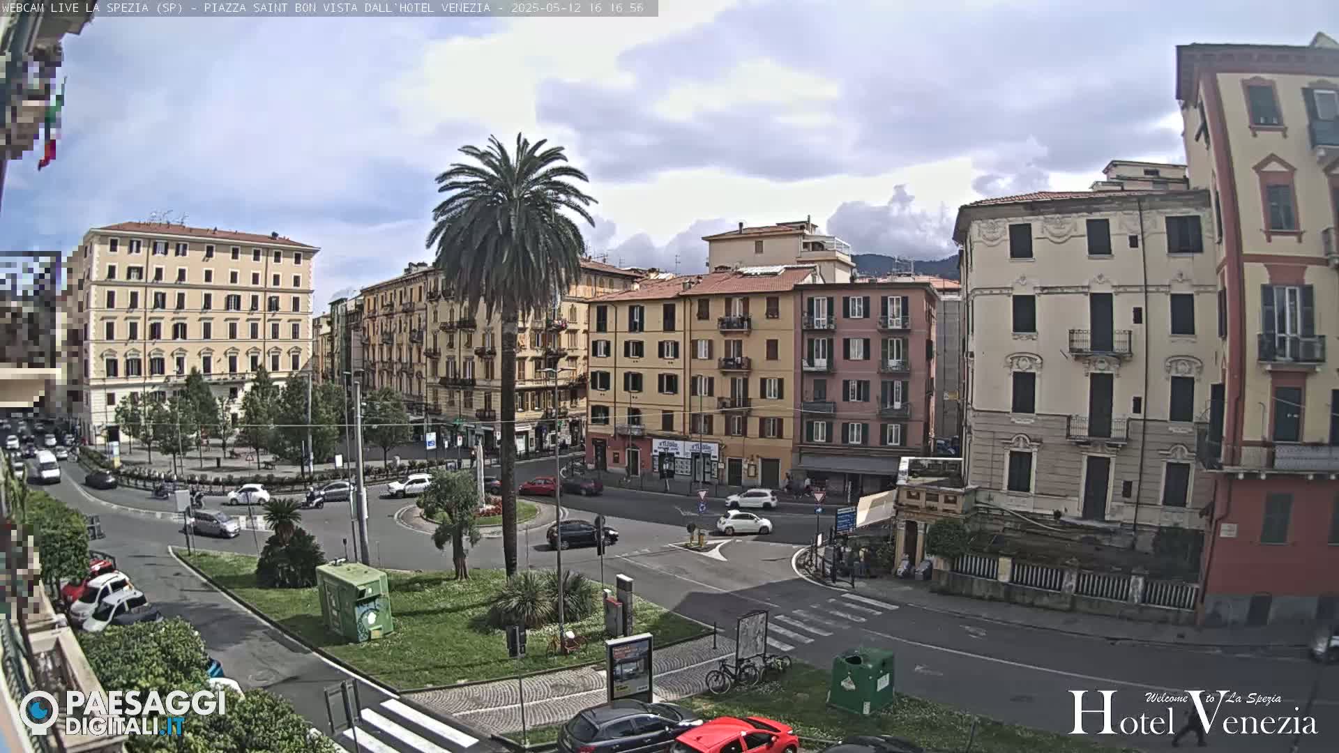 A partly cloudy day reveals a city square with several multi-story buildings, palm trees, and cars, all viewed from an elevated perspective.