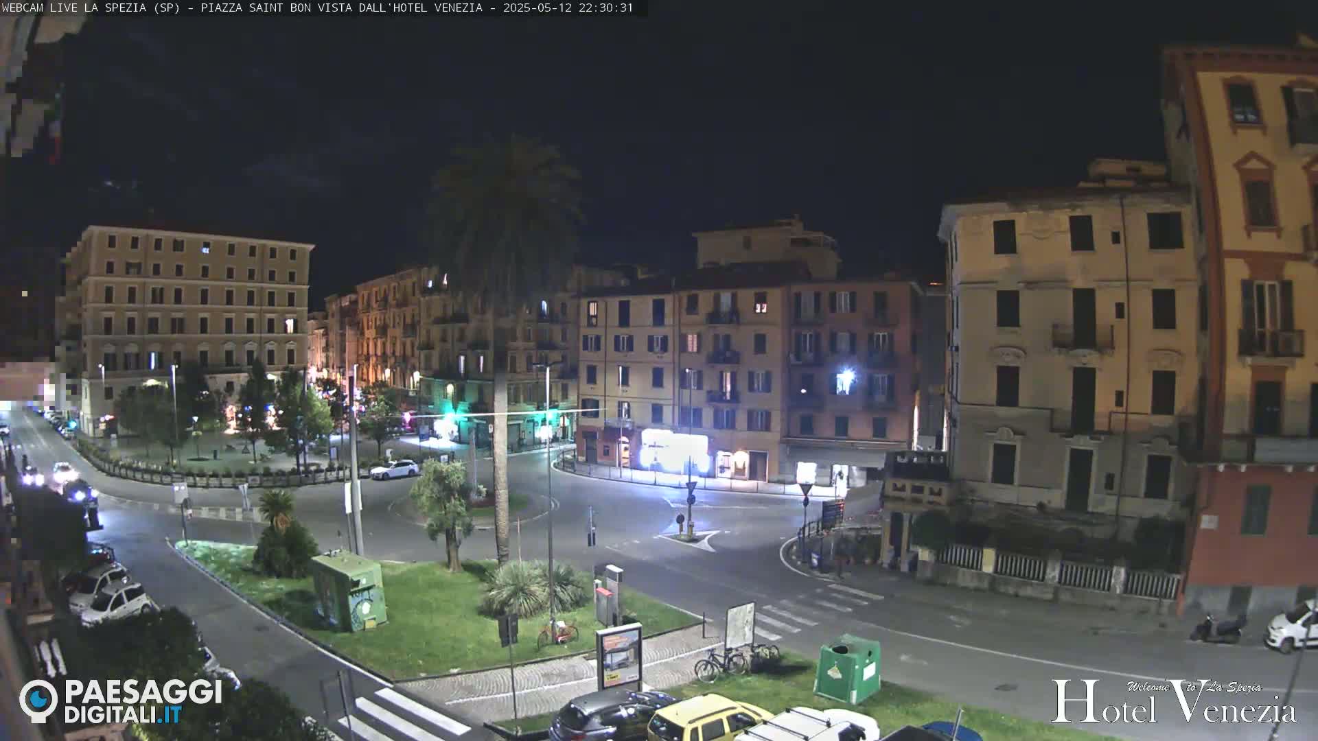 A nighttime, city-square scene shows several multi-story buildings surrounding a circular roadway with sparsely parked cars, a lone palm tree, and minimal pedestrian activity.