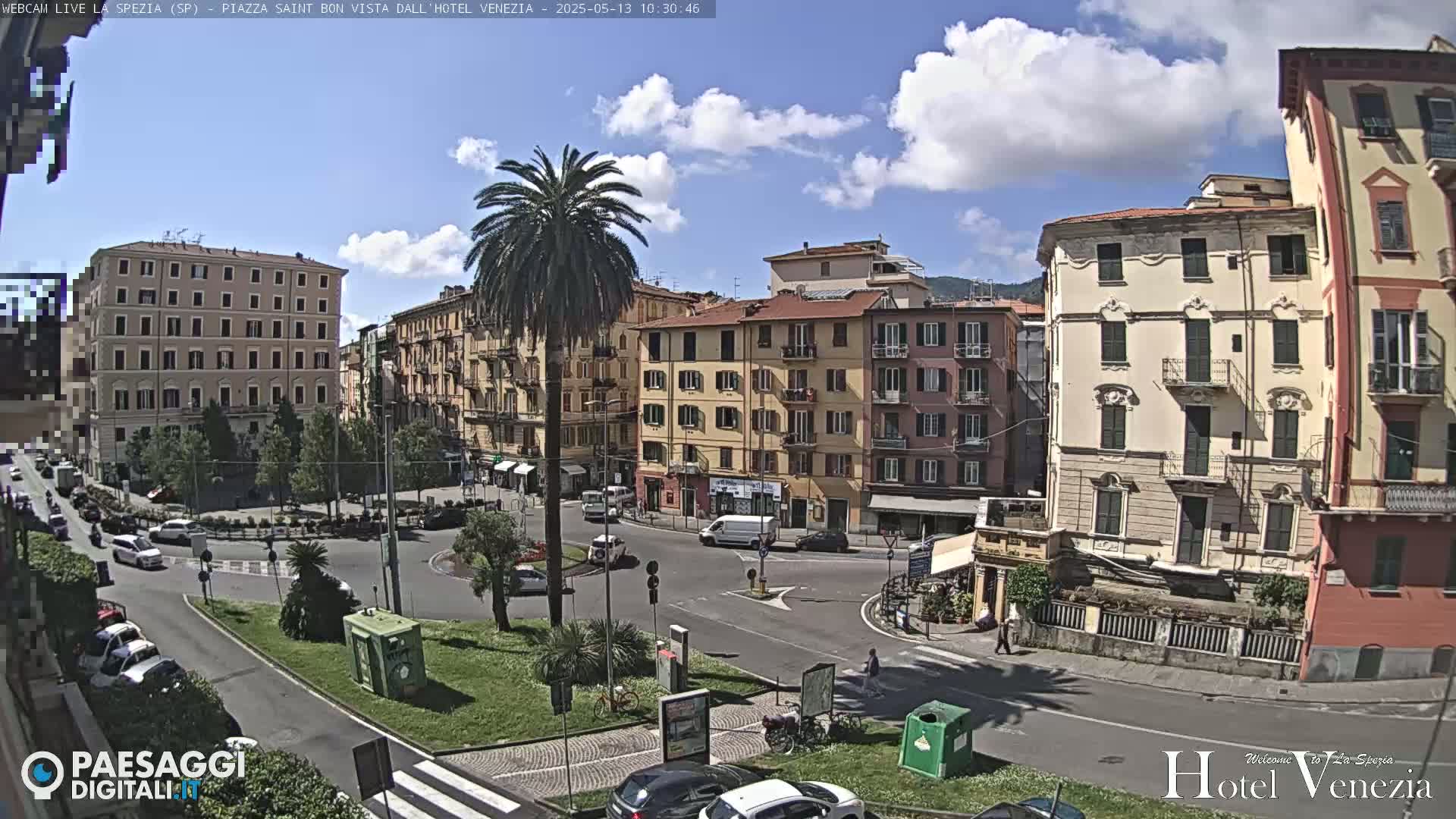 A sunny, partly cloudy day reveals a European city square with multi-story buildings, palm trees, cars, and pedestrians.
