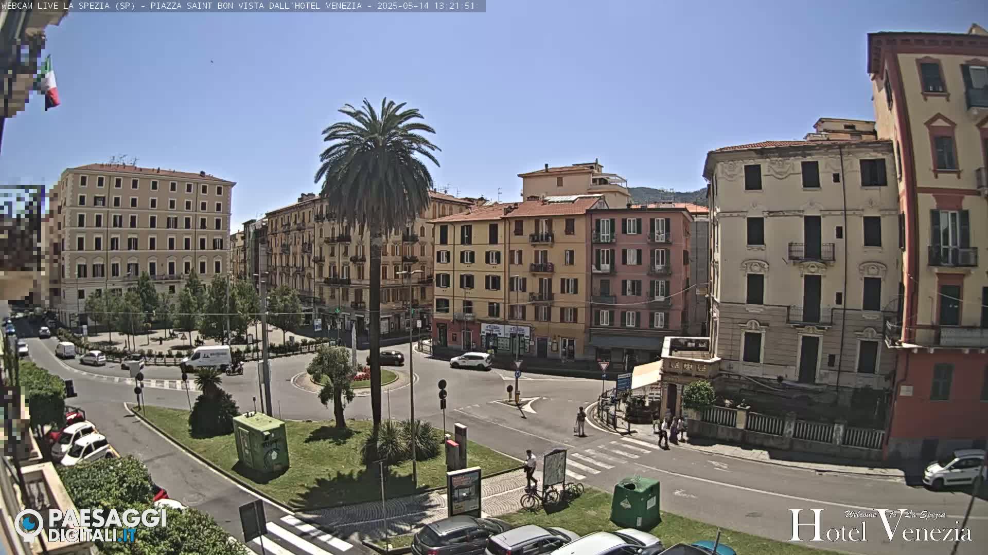 A sunny day reveals a town square with several multi-story buildings, cars, and a few pedestrians; a tall palm tree stands in the center of the square.