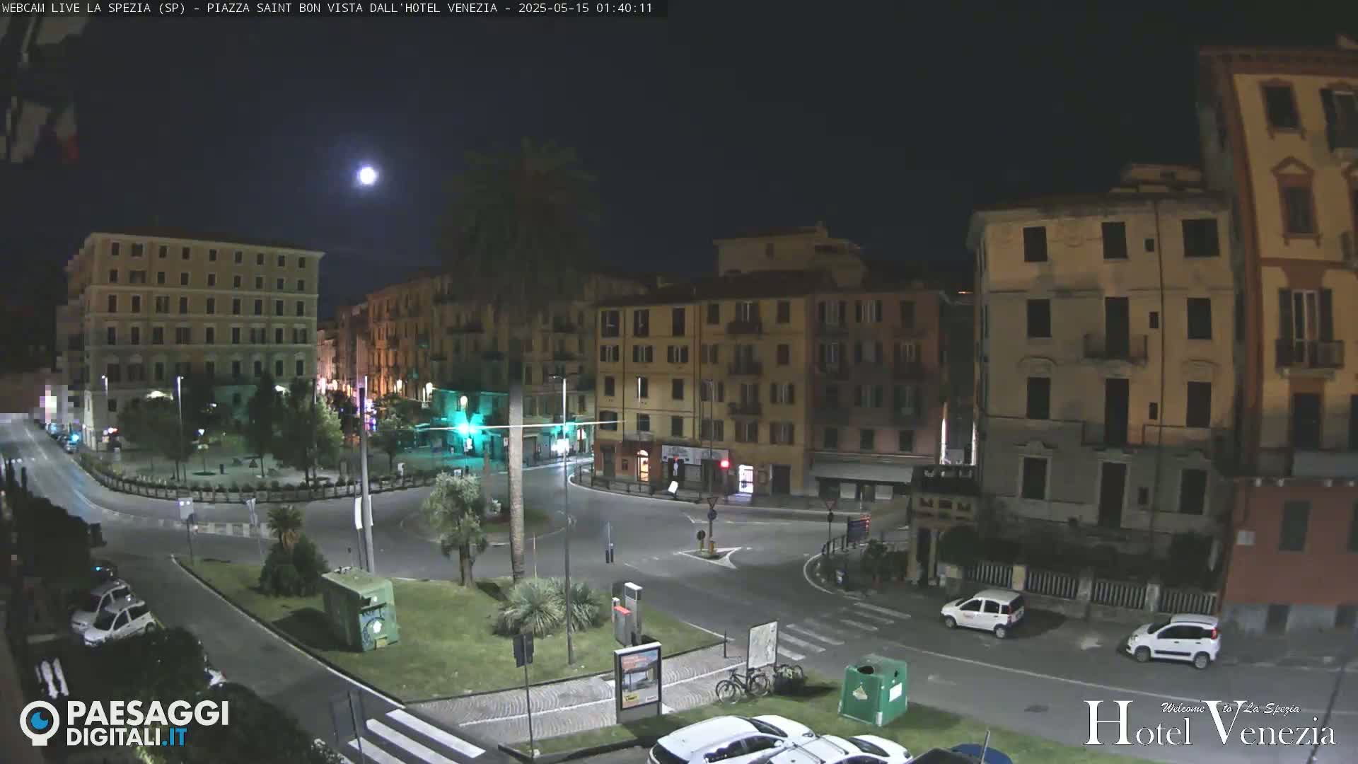 A nighttime view of a town square with several multi-story buildings, a few cars, and a palm tree under a mostly clear night sky with a visible moon.