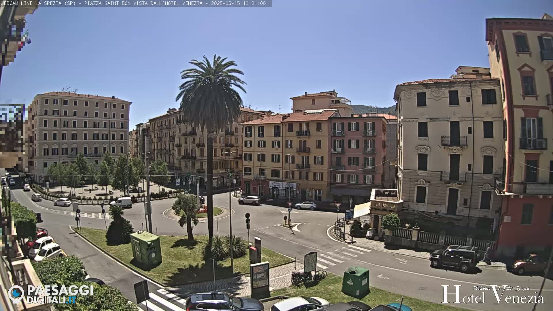 A sunny day in a European city square shows multi-story buildings surrounding a traffic circle with cars and a central, landscaped area featuring a tall palm tree.