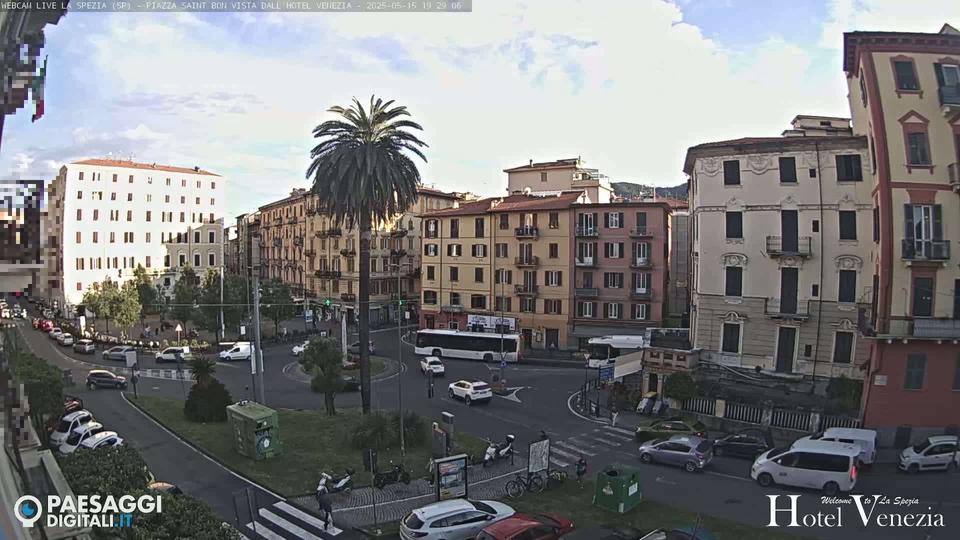 A partly cloudy daytime scene shows a city square with several multi-story buildings, cars, and a palm tree in the center, all surrounded by roadways.