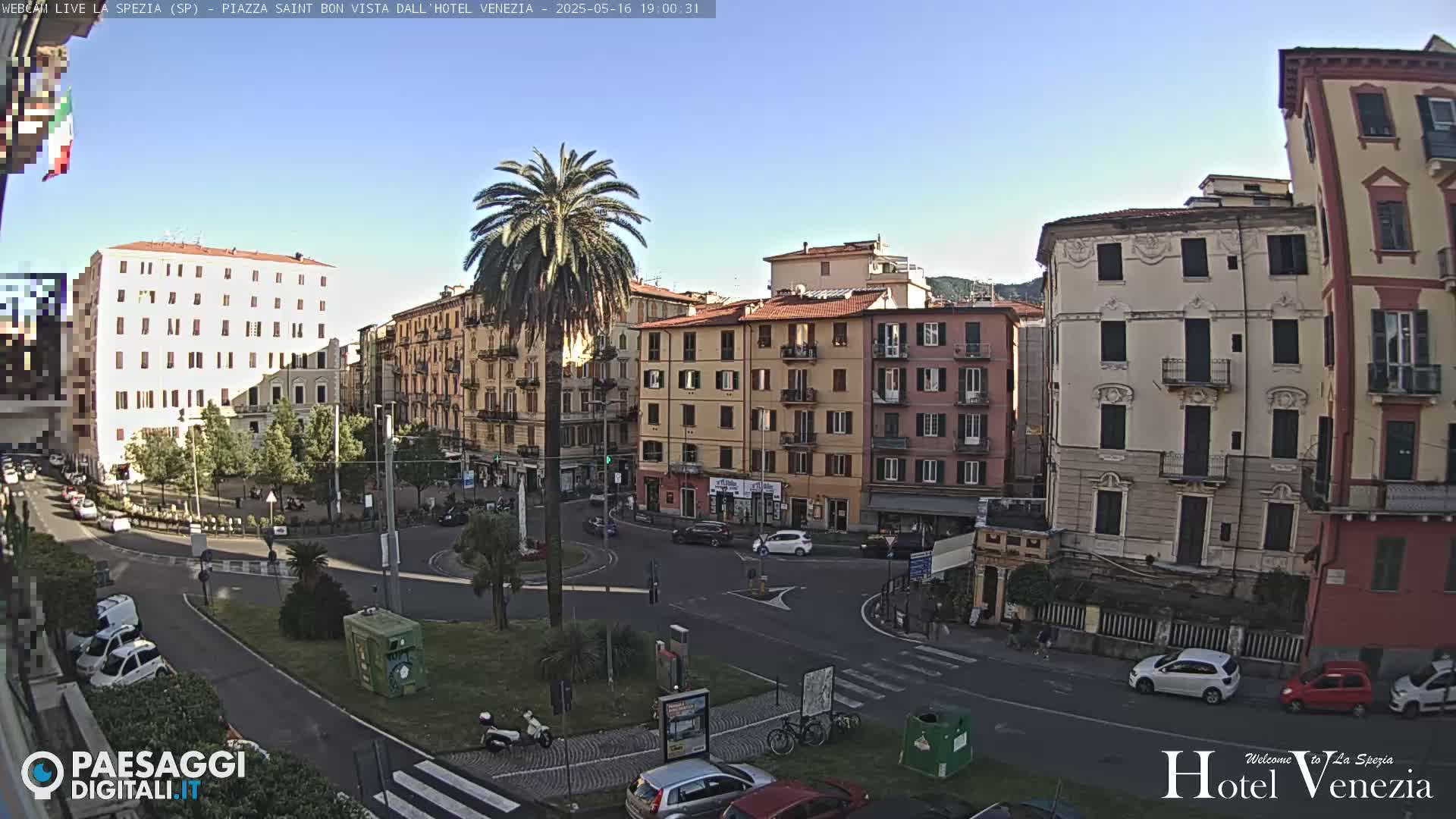 A sunny day in a European city square, showing several multi-story buildings surrounding a traffic circle with cars and pedestrians.