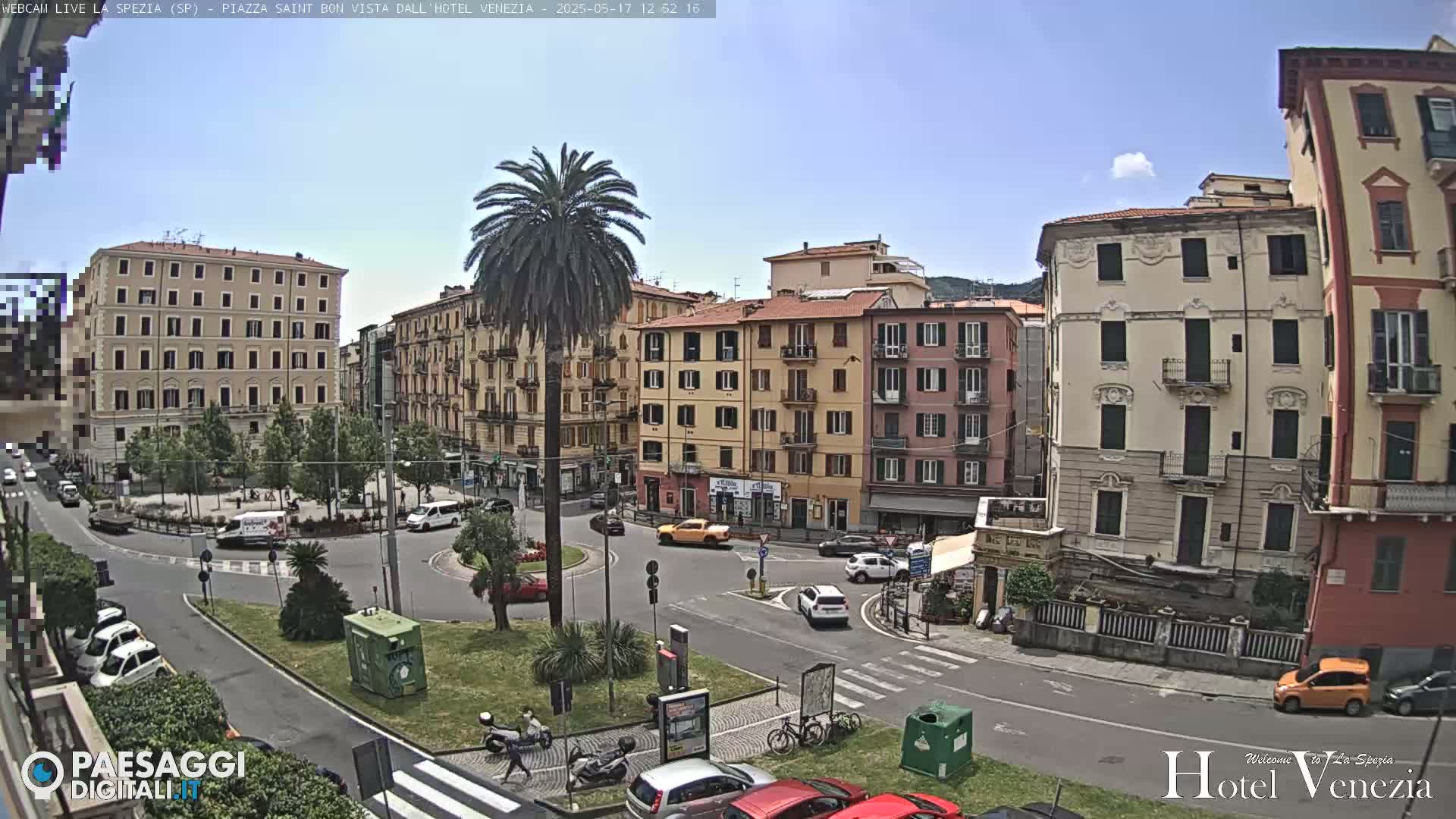 A sunny day reveals a European city square with several multi-story buildings surrounding a small roundabout with cars and a central palm tree.
