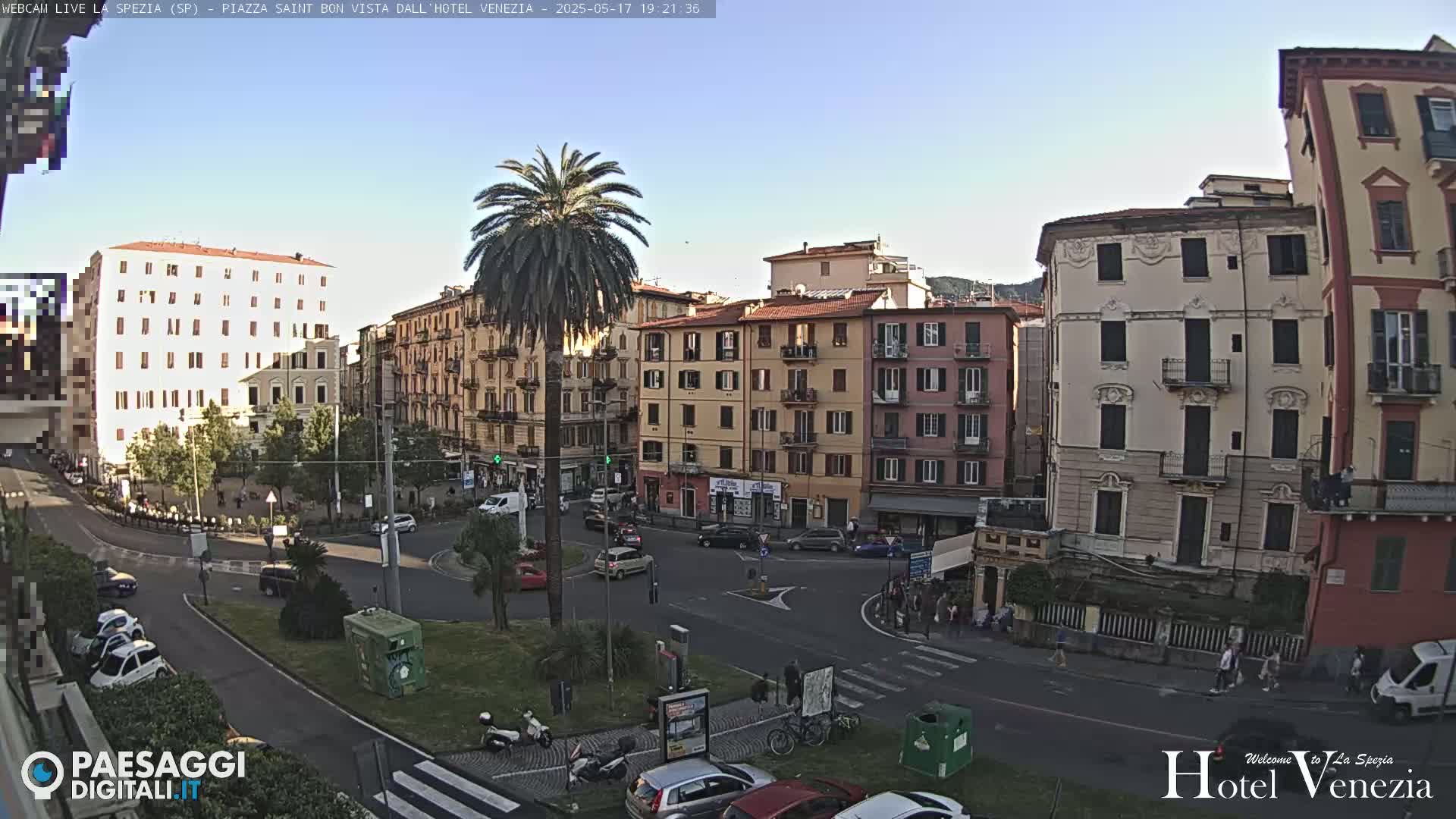 A sunny, late afternoon view of a European town square, showing several multi-story buildings, cars, pedestrians, and a lone palm tree in the center.
