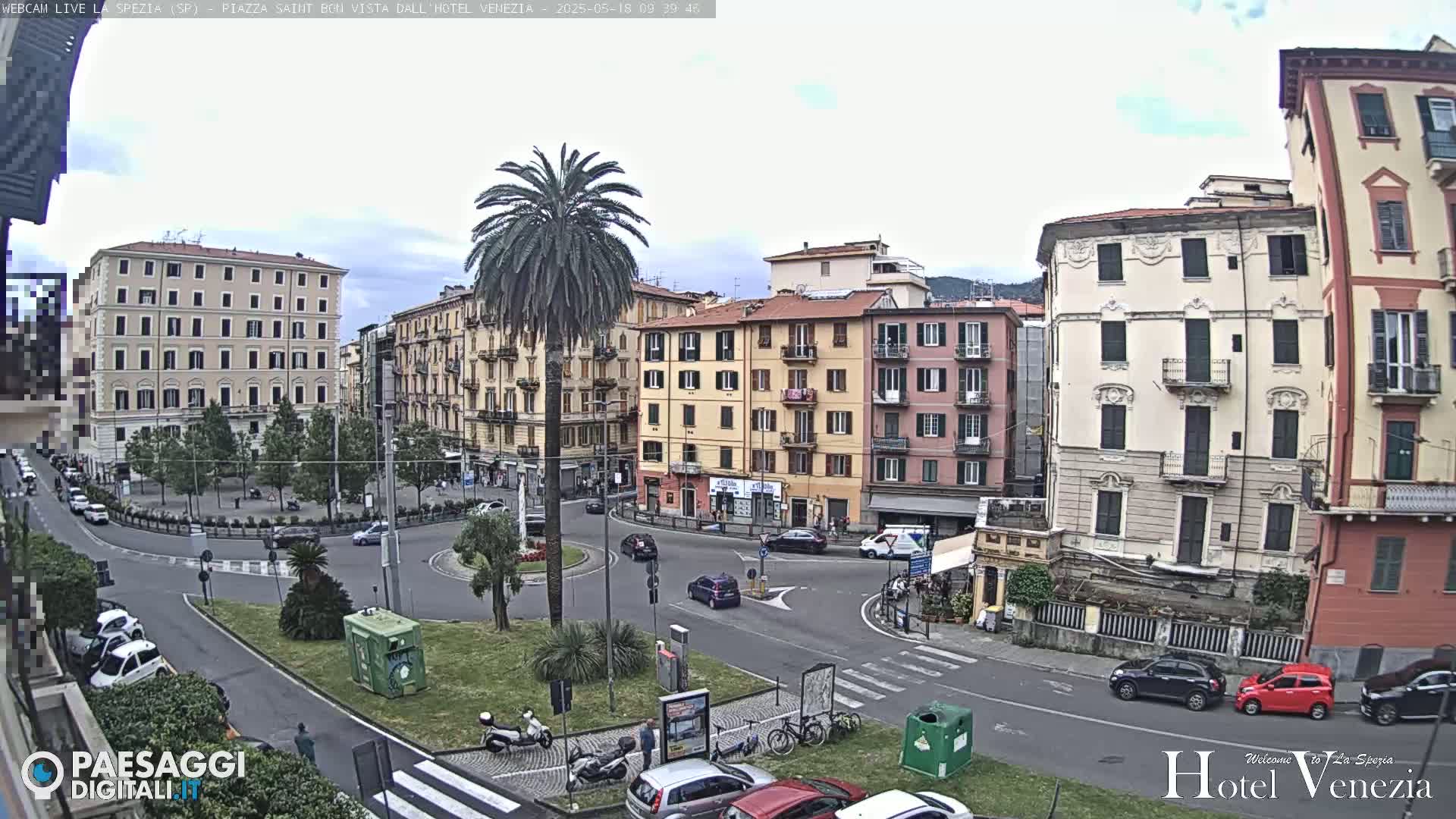 A mostly cloudy day reveals a European city square with several multi-story buildings surrounding a roundabout with cars and pedestrians, and a tall palm tree in the center.