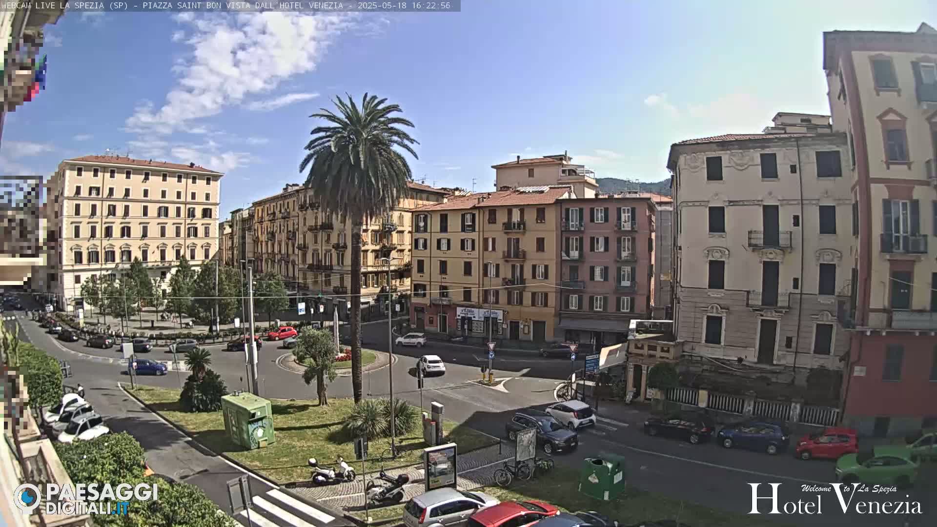 A sunny day in a European city square, showing several multi-story buildings surrounding a roundabout with cars and a few pedestrians.