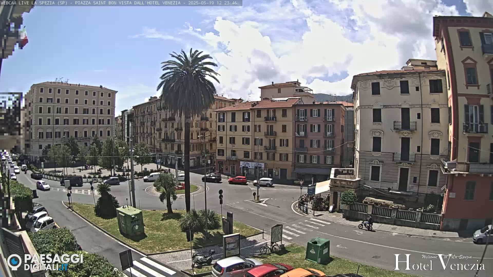 A sunny day in a European city square shows several multi-story buildings surrounding a roundabout with cars and a palm tree in the center.