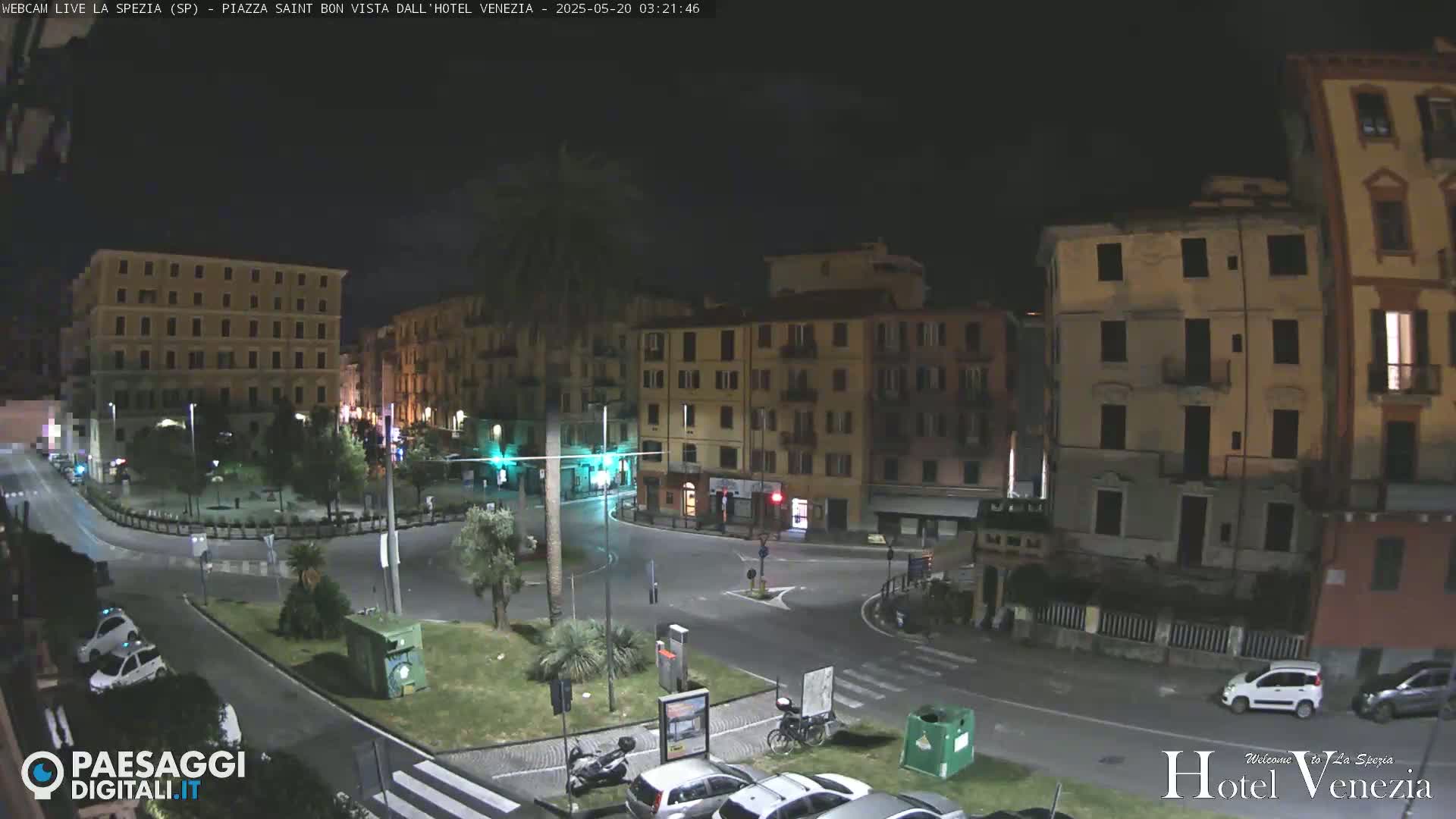 A nighttime view of a town square with several multi-story buildings surrounding a circular roadway, with a few cars and a scooter parked.