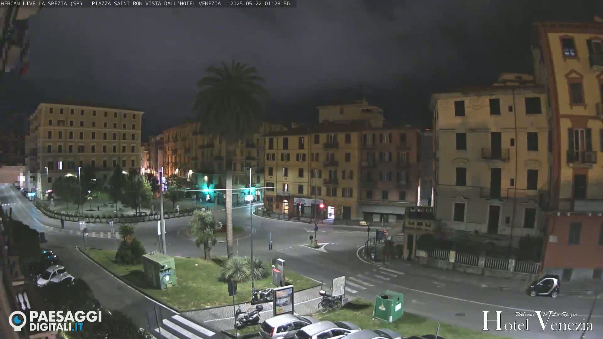 A nighttime, dimly lit city square with several multi-story buildings surrounding a circular roadway, featuring sparse vehicle traffic and a lone palm tree.