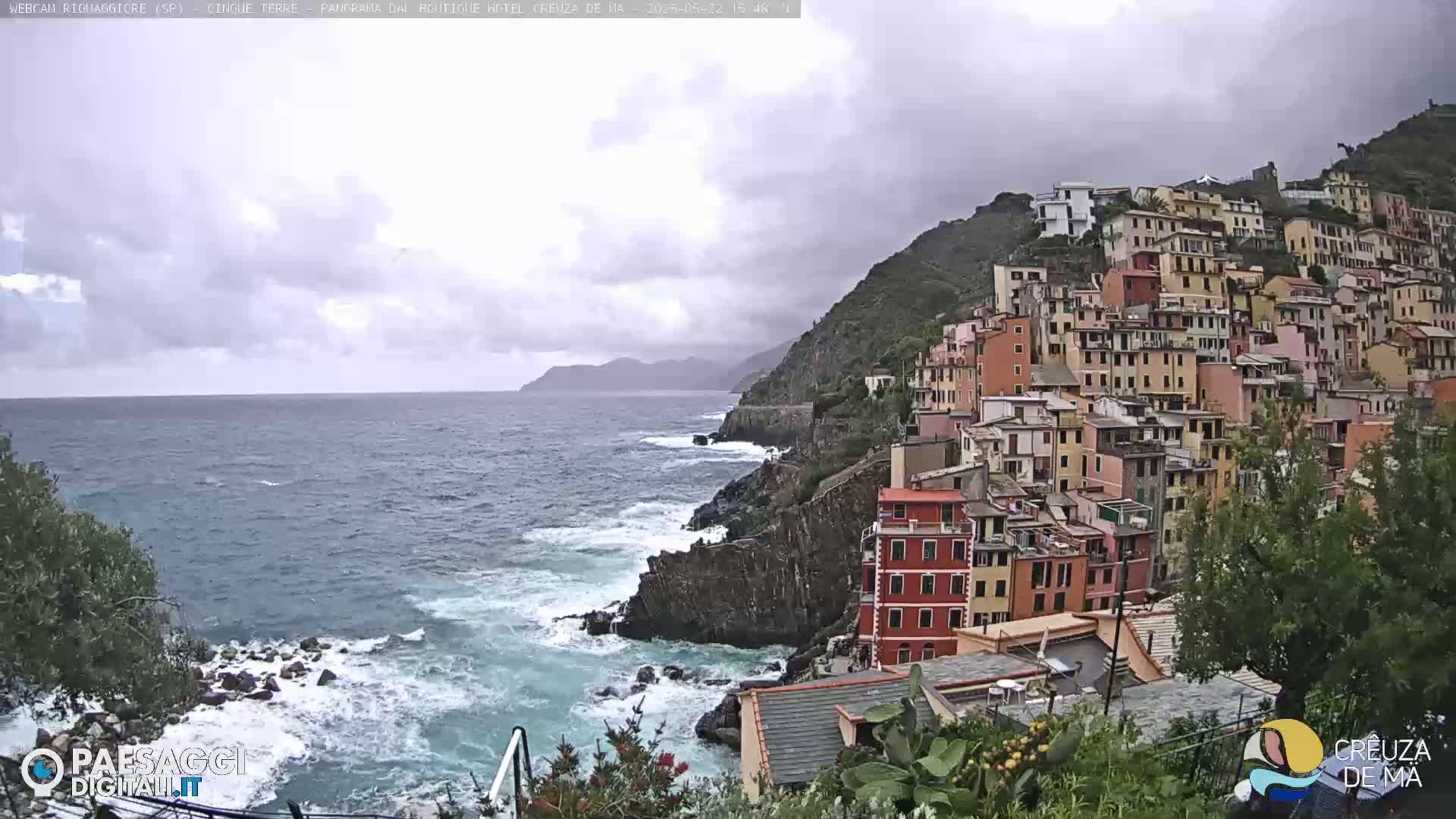 A seaside village of colorful buildings clings to a steep cliff overlooking a rough sea under a cloudy sky.