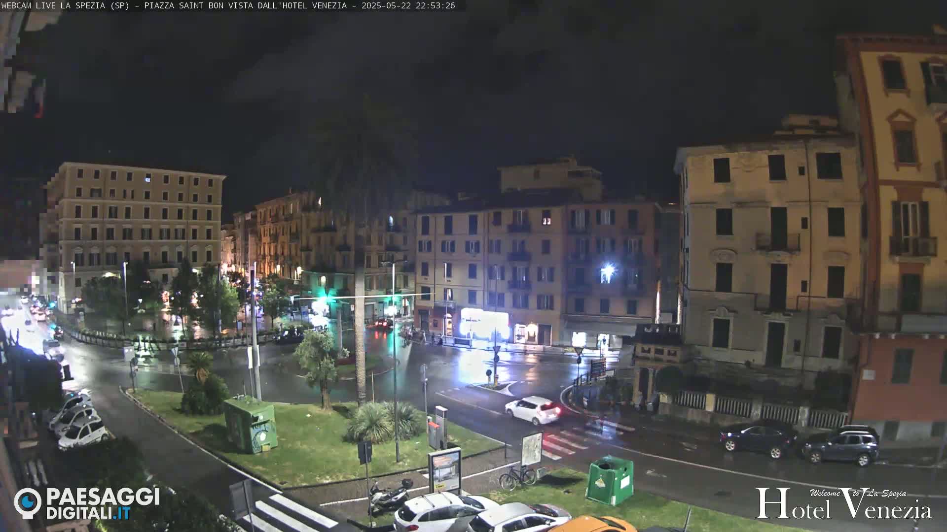 A rain-slicked nighttime city square, surrounded by multi-story buildings, with cars and a few people visible.