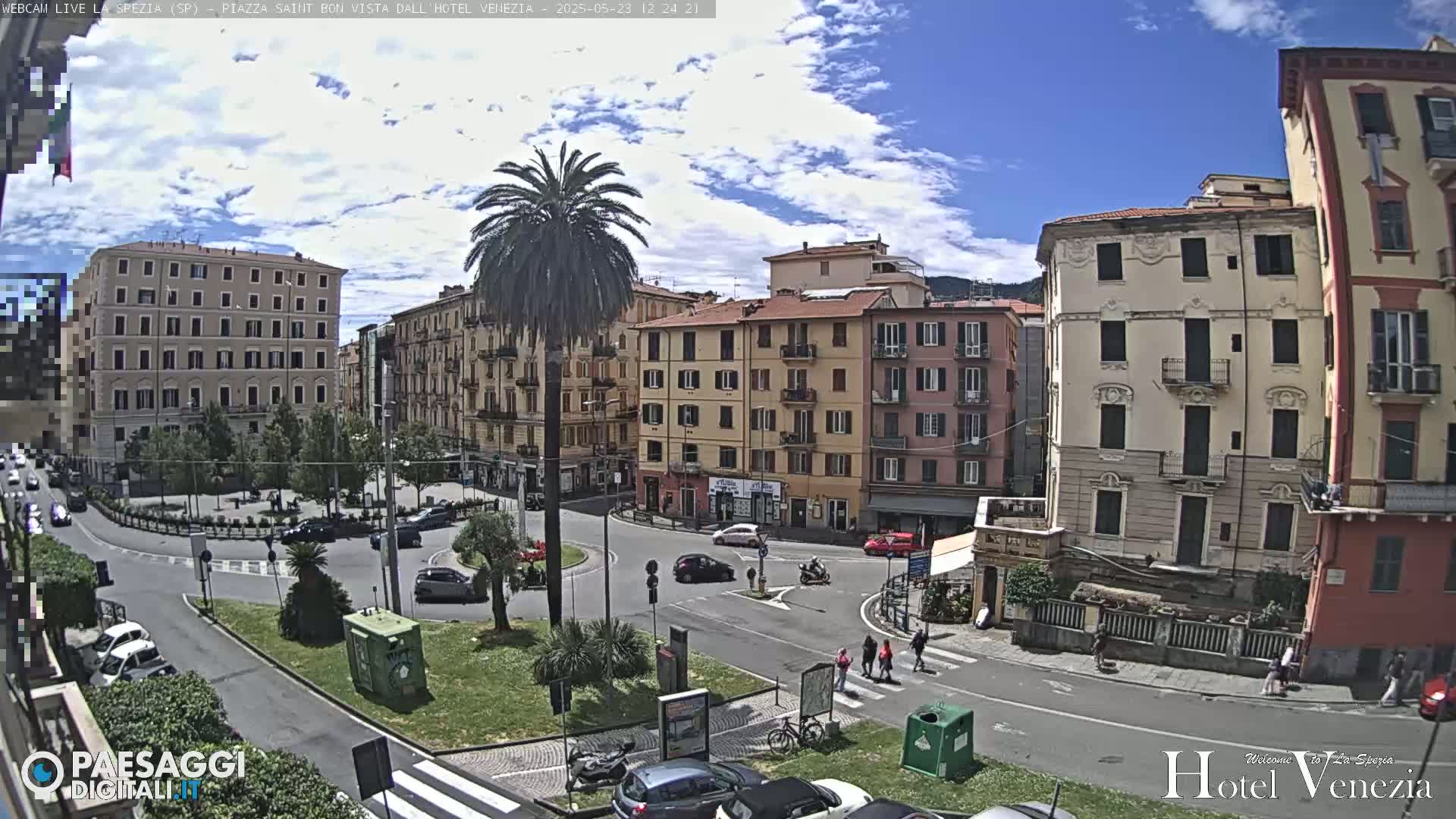 A sunny day in a European town square, showing several multi-story buildings surrounding a roundabout with cars and pedestrians.