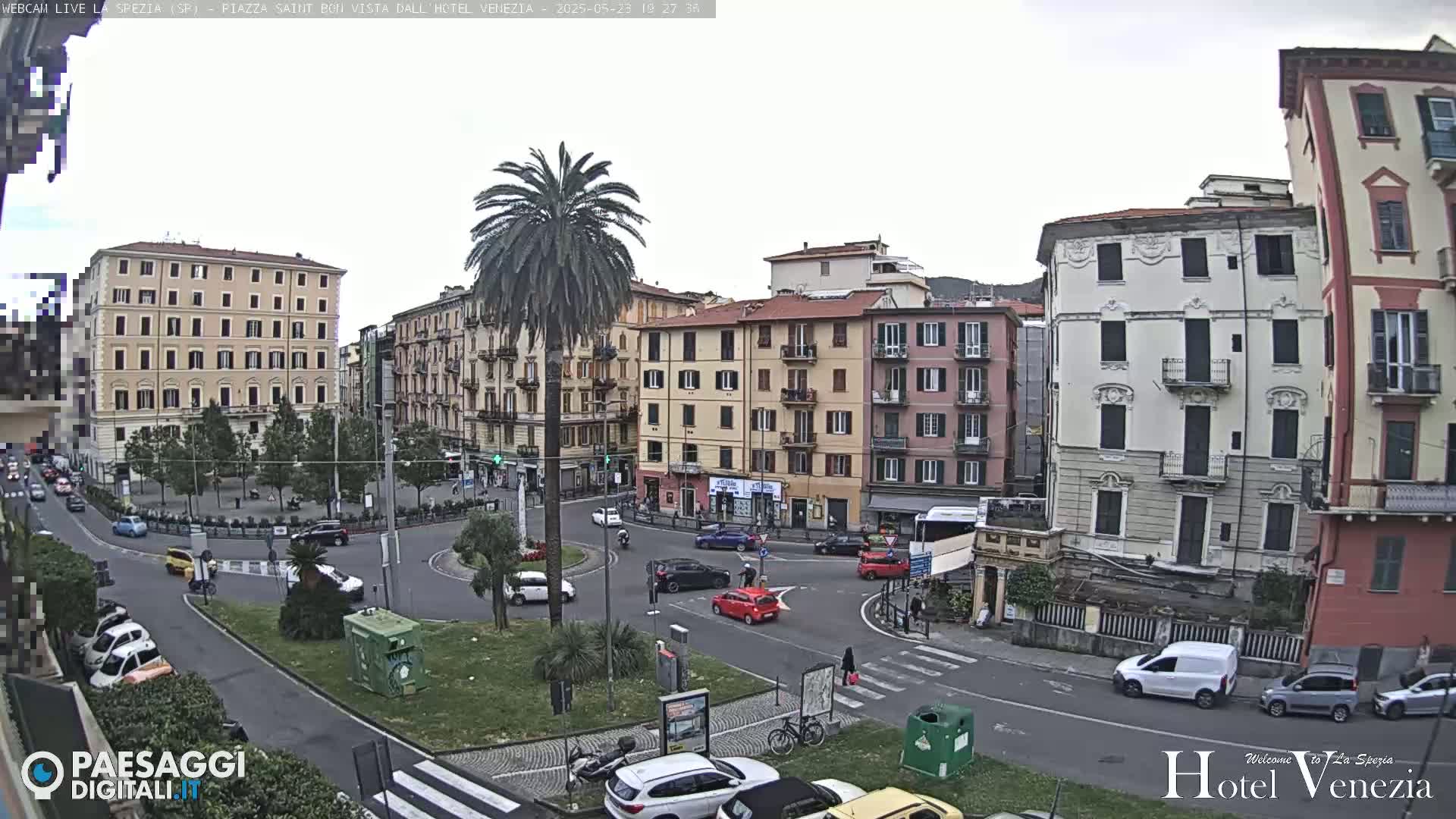 A city square with several multi-story buildings, some cars, a palm tree, and overcast skies.