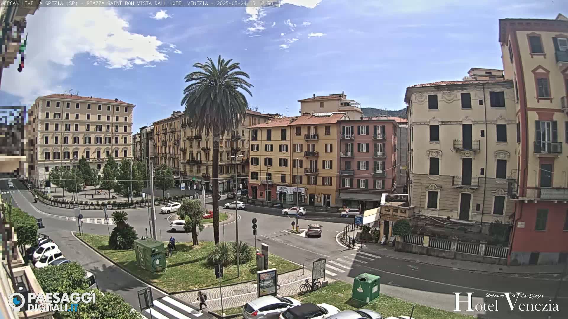 A sunny, partly cloudy day overlooks a European-style plaza with several multi-story buildings, a central roundabout with cars, and a tall palm tree.