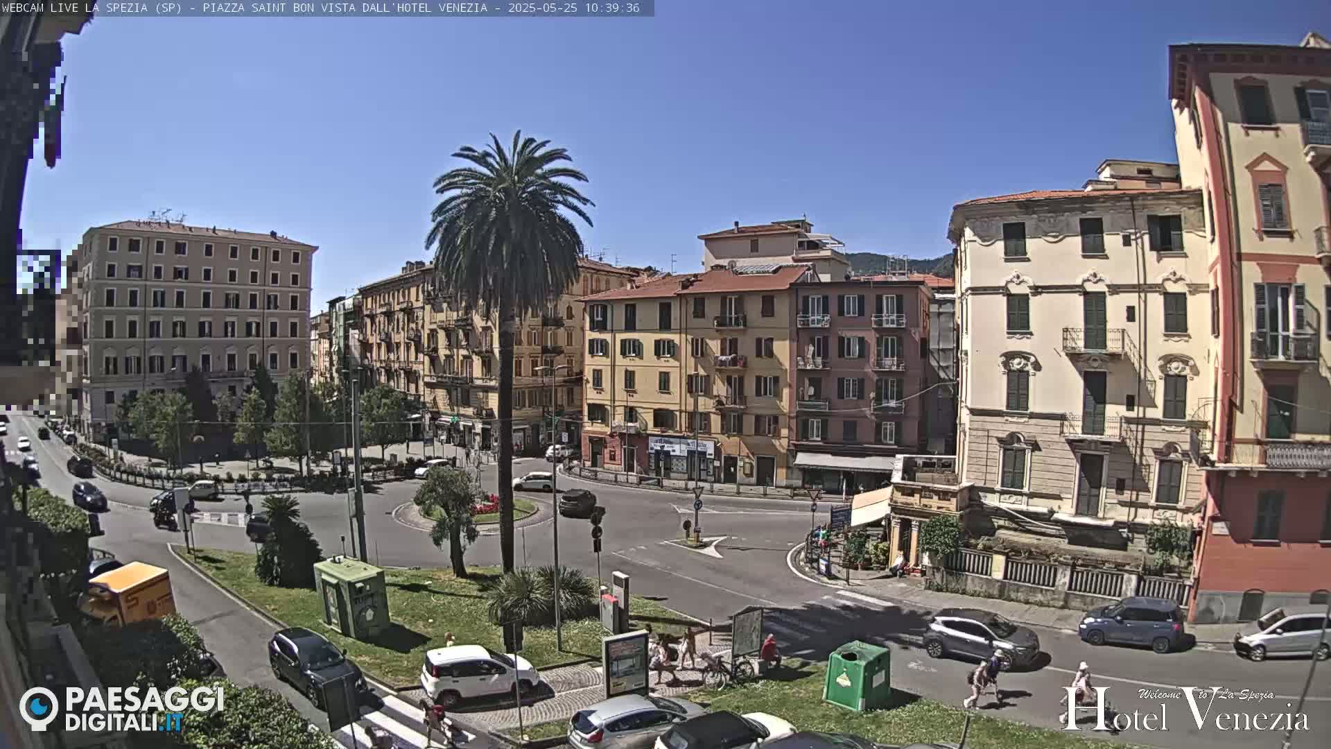 A sunny day in a European city square, showing several multi-story buildings and cars around a roundabout with pedestrians and palm trees.