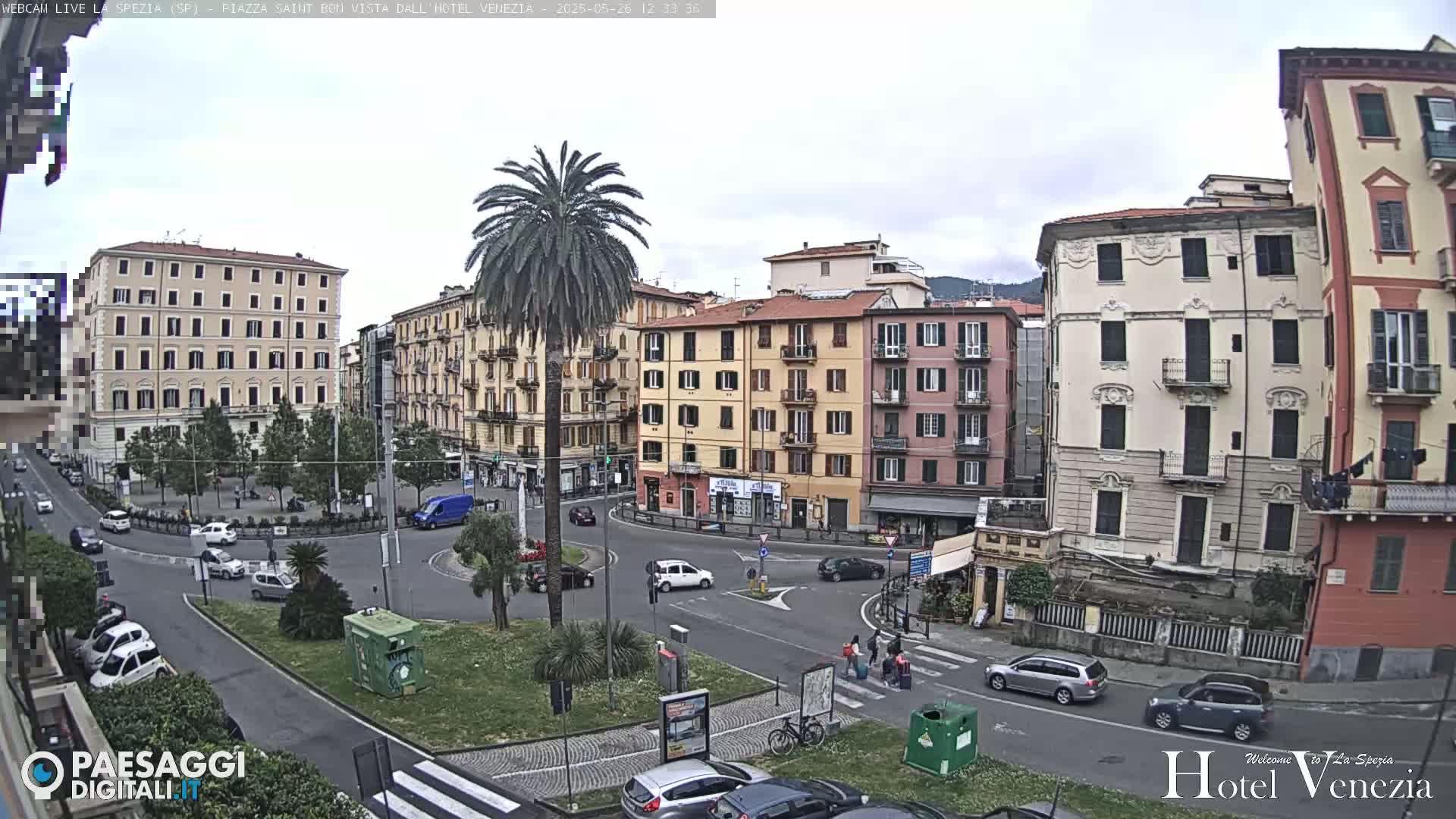 A mostly cloudy day reveals a city square with several multi-story buildings, cars, and a palm tree, surrounded by green landscaping.