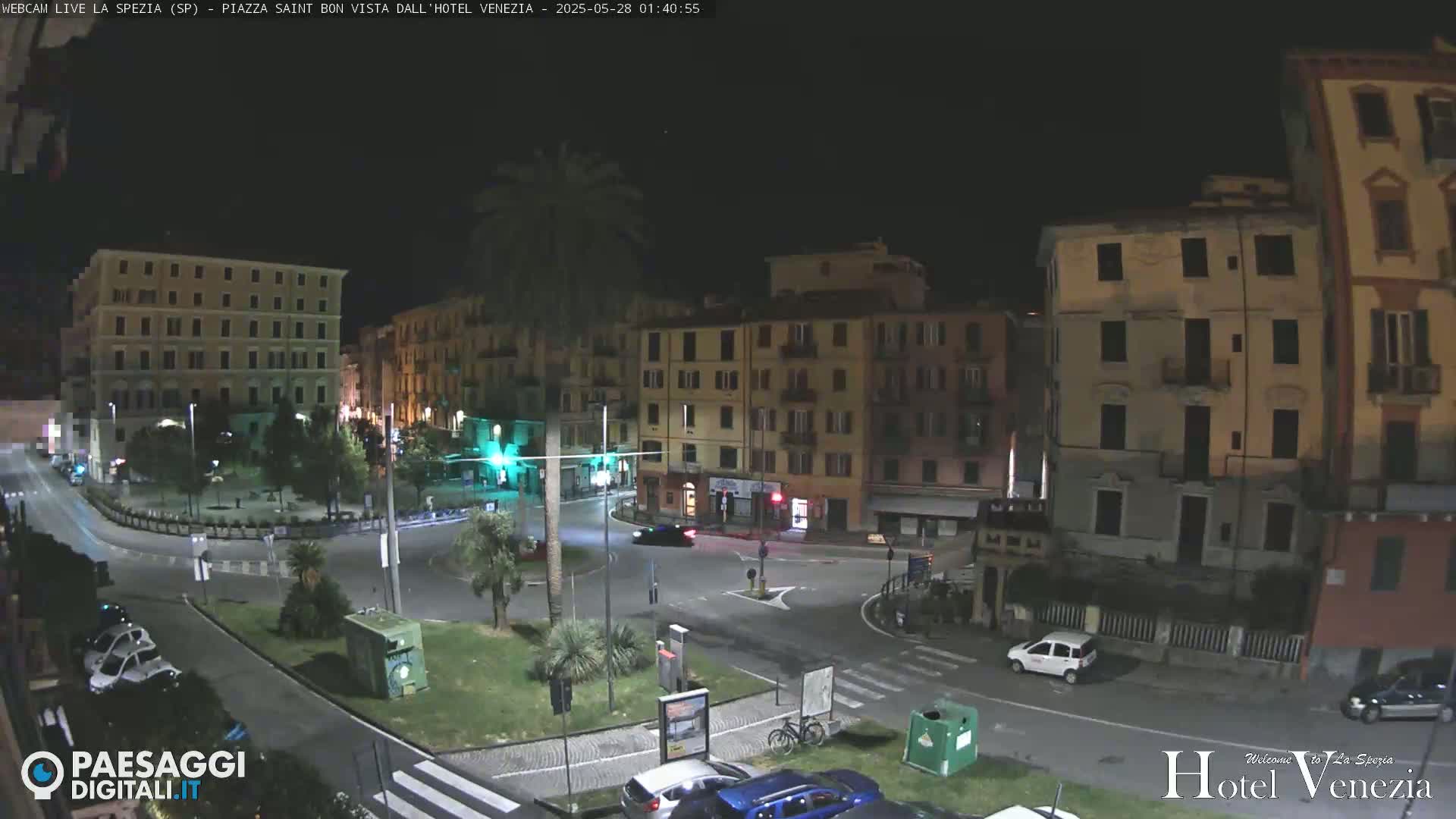 A nighttime view of a city square with several multi-story buildings surrounding a traffic circle, illuminated by streetlights.