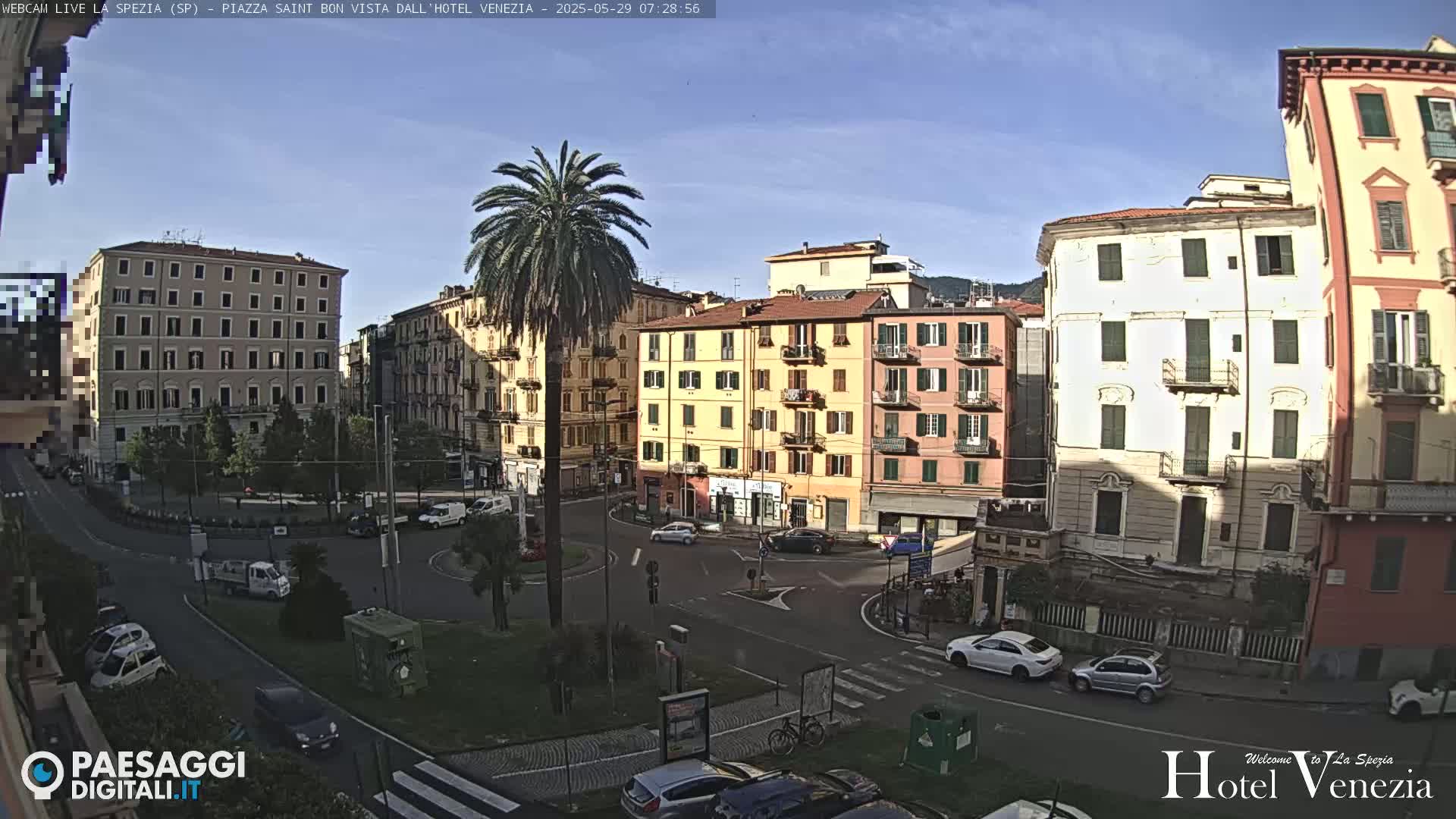 A sunny day overlooks a city square with several multi-story buildings, palm trees, and cars parked and driving around the square.