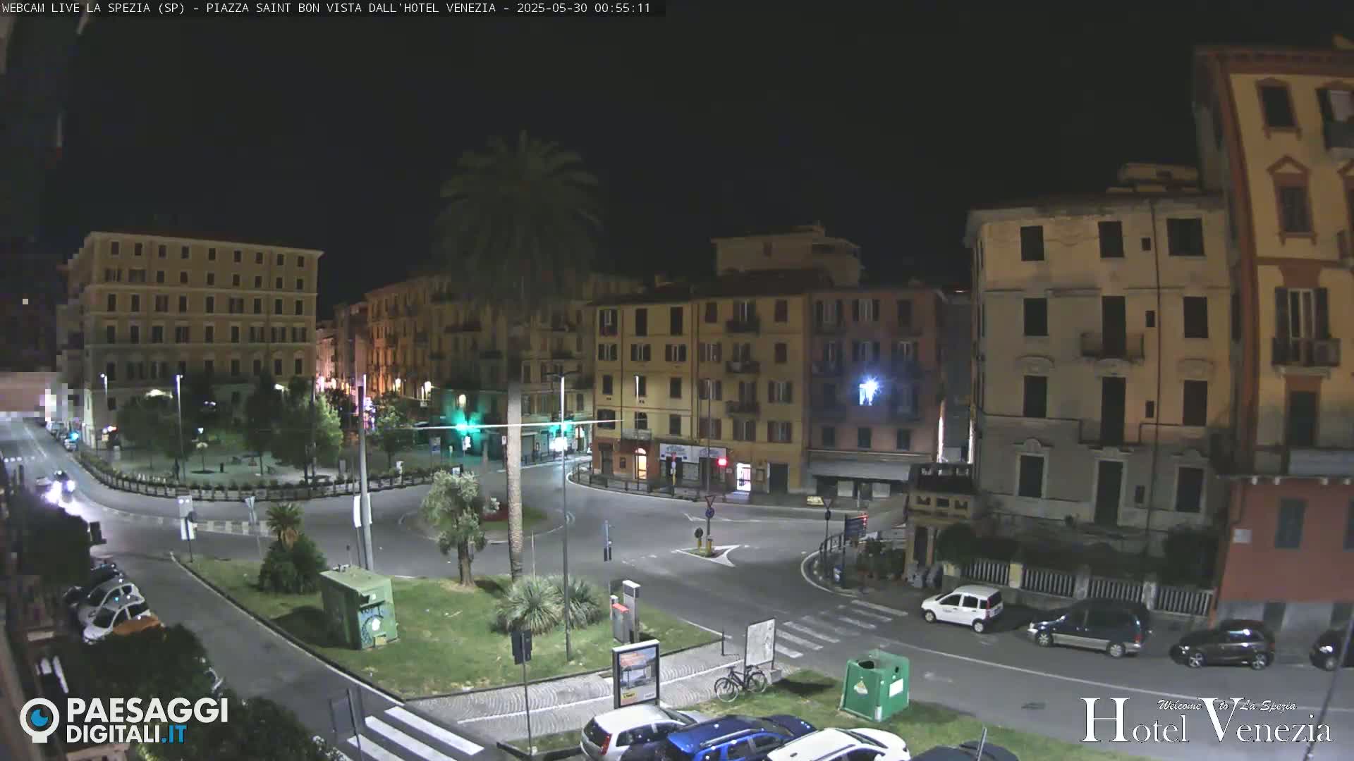 A nighttime, high-angle view of a mostly empty town square in Italy, surrounded by multi-story buildings, with a few cars parked and driving.