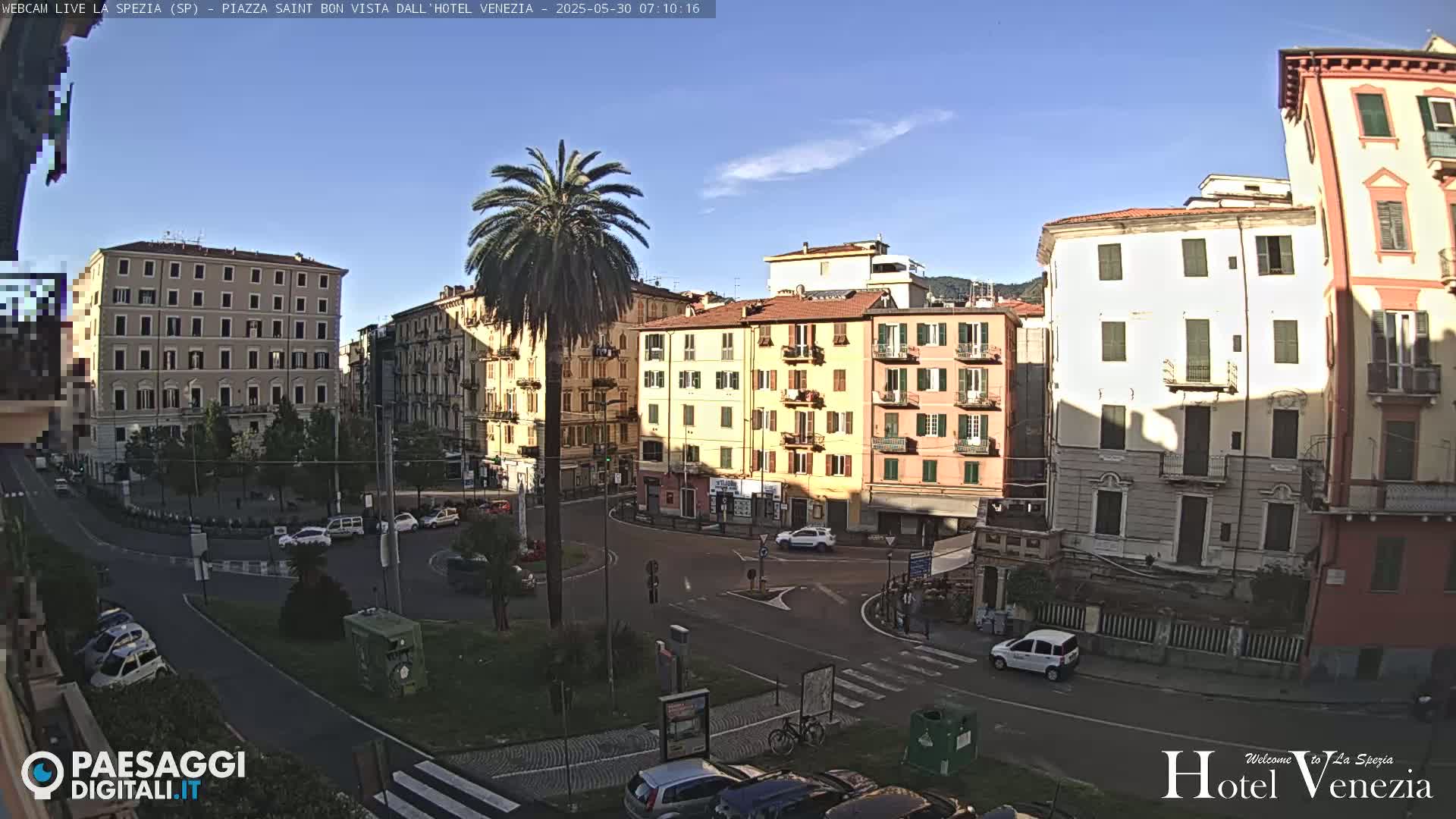 A sunny day reveals a European city square with several multi-story buildings, a palm tree, and a few cars.