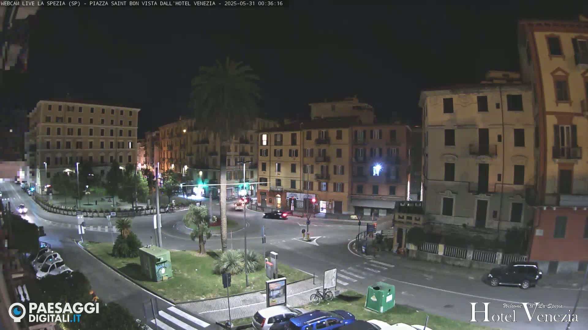 A nighttime view of a city square with several multi-story buildings surrounding a roundabout, with a few cars and a bicycle visible.