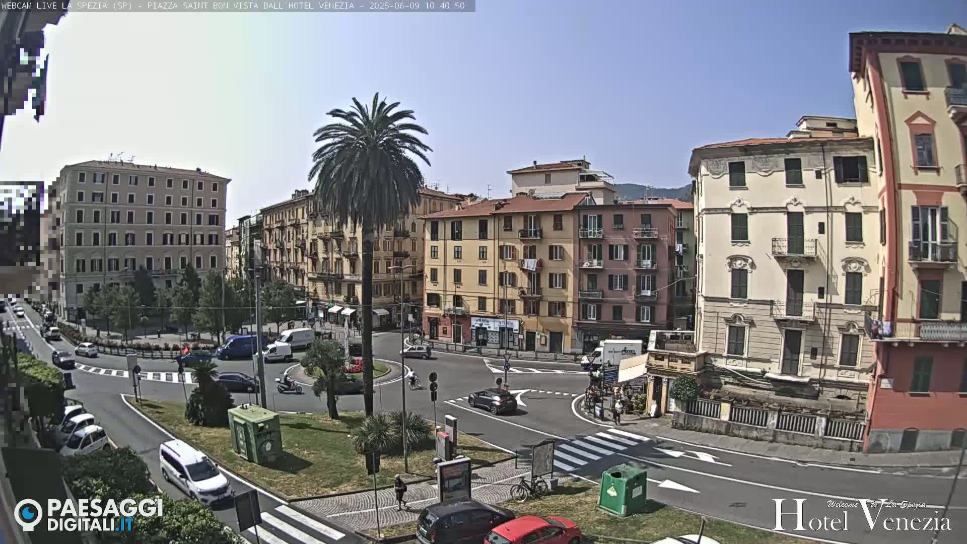 A sunny day reveals a city square with several multi-story buildings, palm trees, and cars, in a roundabout.