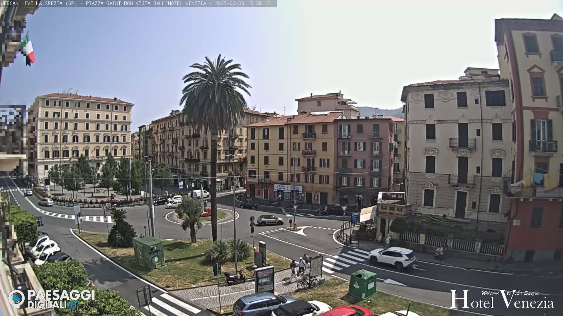A sunny day in a European town square, showing several multi-story buildings surrounding a roundabout with sparse traffic and pedestrians.