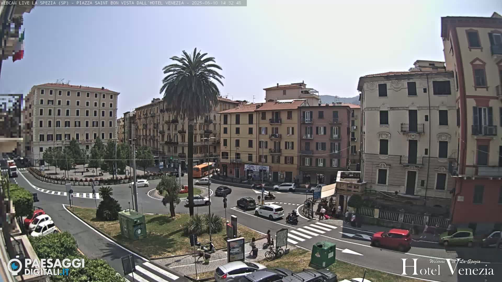 A sunny day in a European town square, showing several multi-story buildings, cars, pedestrians, and a palm tree in the center of a roundabout.