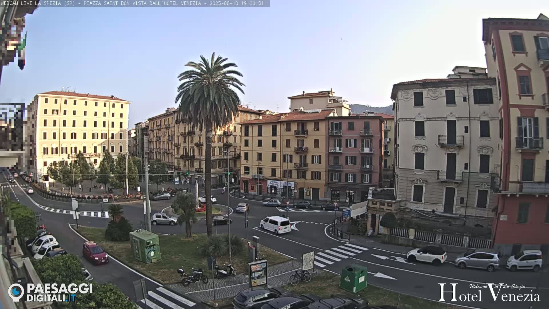 A sunny daytime view of a European city square with several multi-story buildings surrounding a roundabout where cars are driving, with a palm tree in the center.