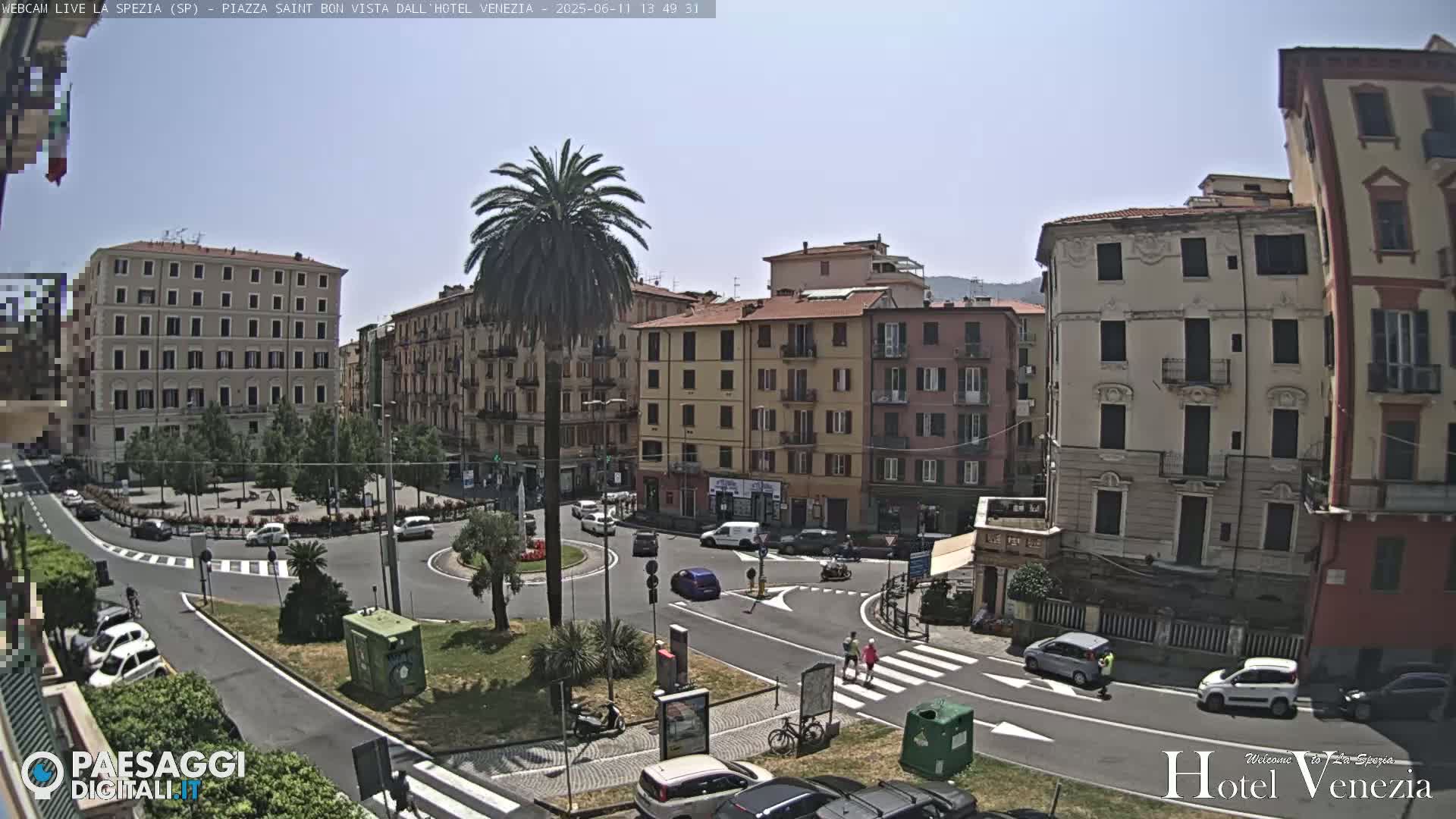 A sunny day overlooking a town square with several multi-story buildings, palm trees, cars, and pedestrians.