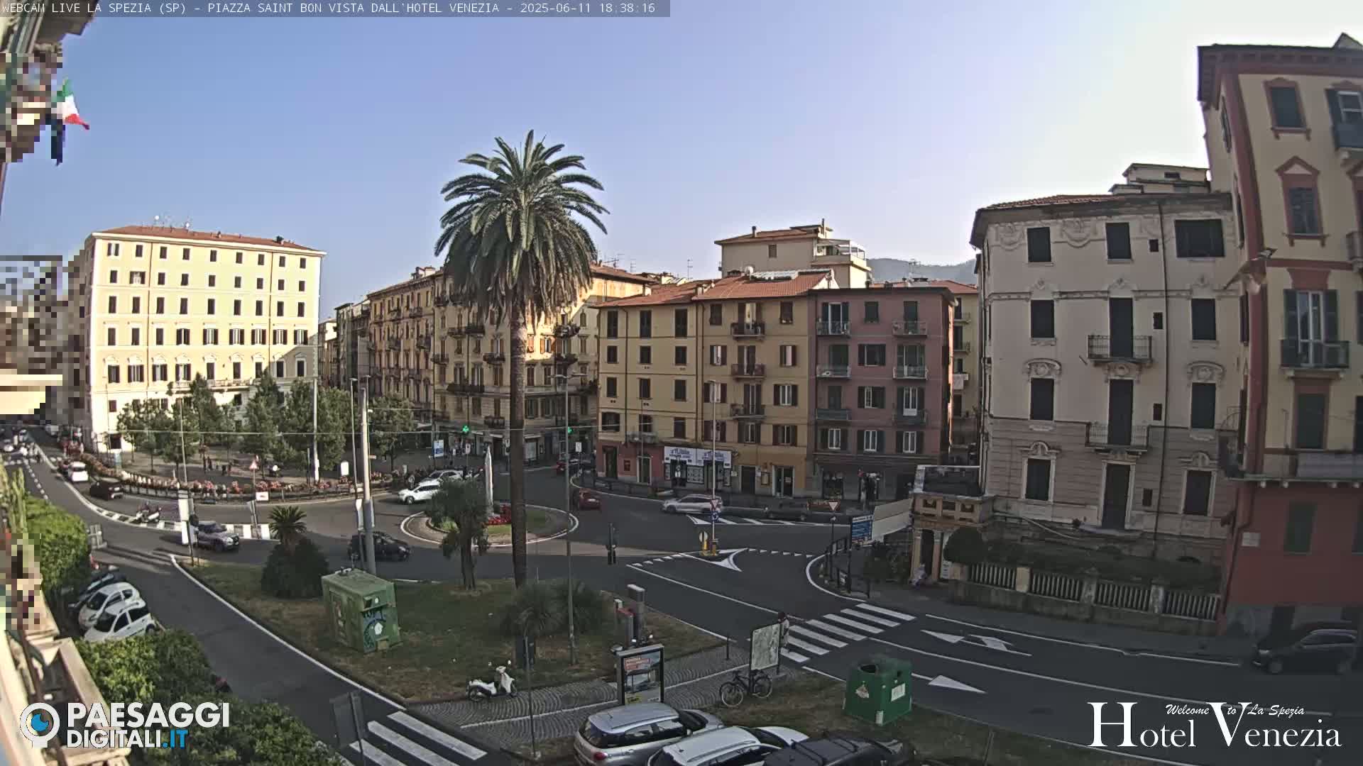 A sunny view of a European city square with multi-story buildings, palm trees, a roundabout, and several vehicles parked and in motion.