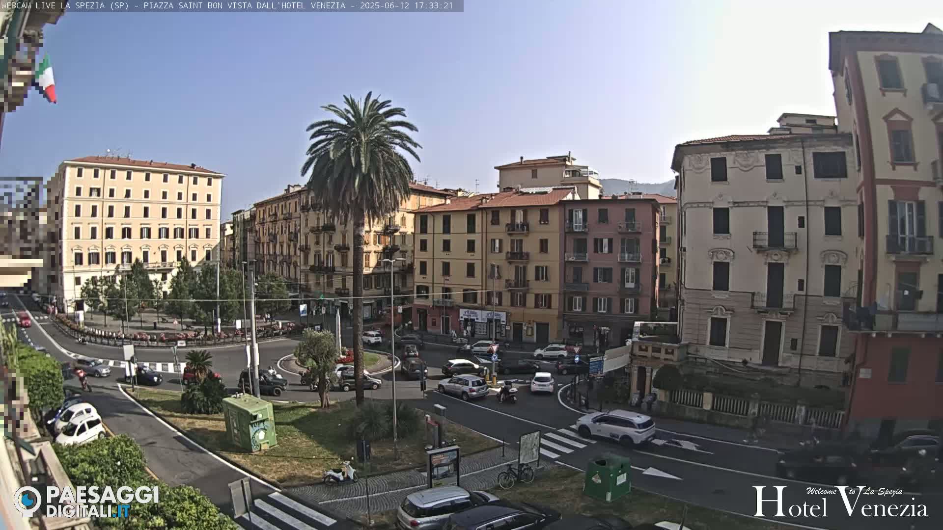 A sunny day overlooking a European city square with several multi-story buildings, a roundabout with cars, and a tall palm tree in the center.