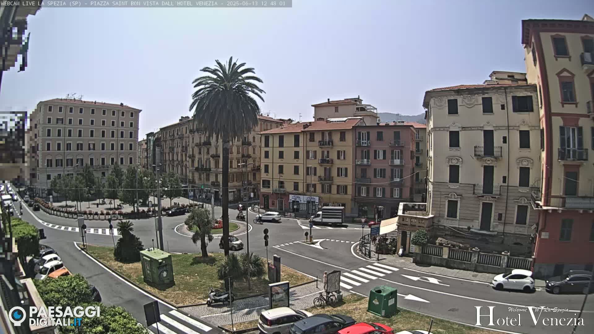 A sunny day overlooking a European town square with several multi-story buildings, a roundabout, parked cars, and a tall palm tree in the center.