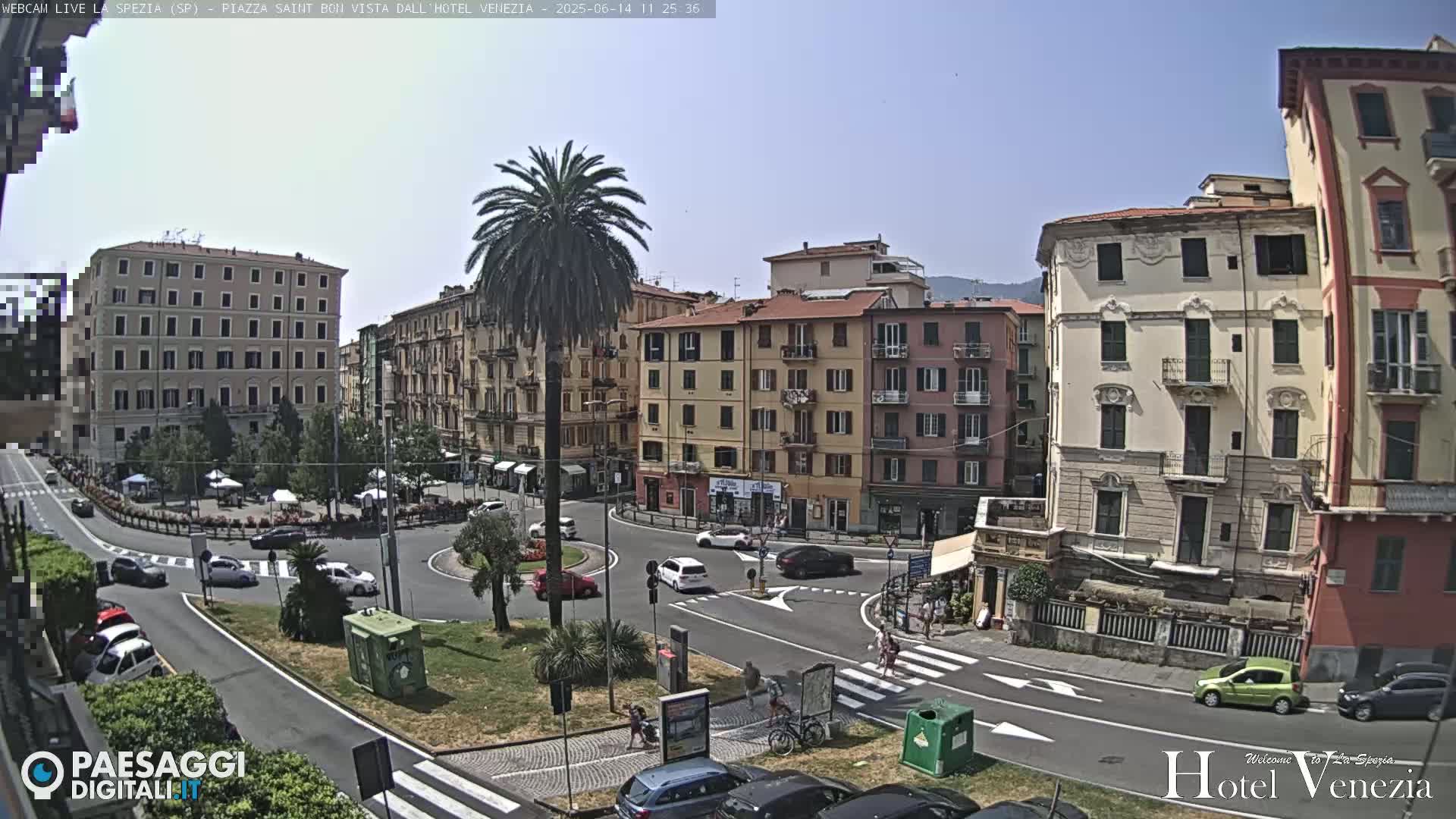 A sunny, outdoor view of a European town square with several multi-story buildings, cars, and pedestrians, centered around a circular traffic island with a palm tree.