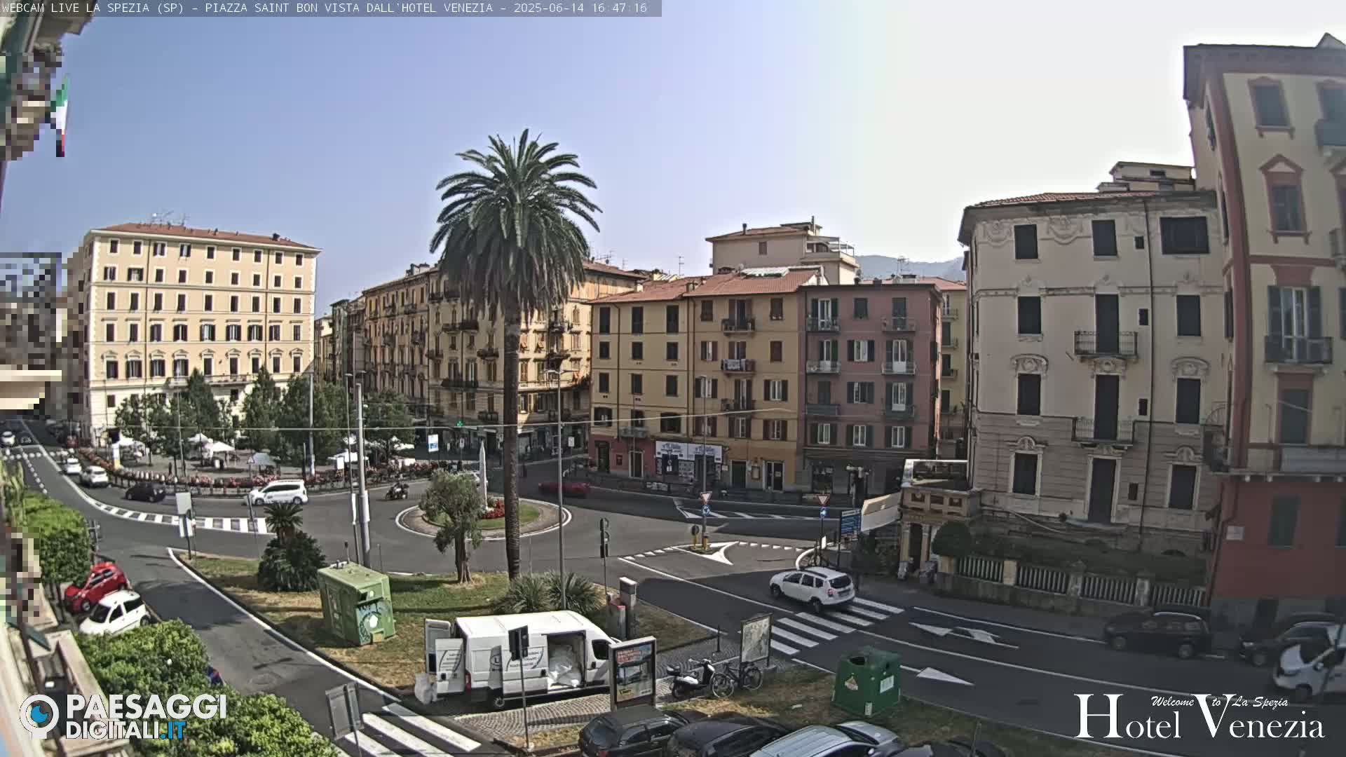 A sunny day overlooks a European town square with several multi-story buildings surrounding a traffic circle with sparse vehicle traffic.