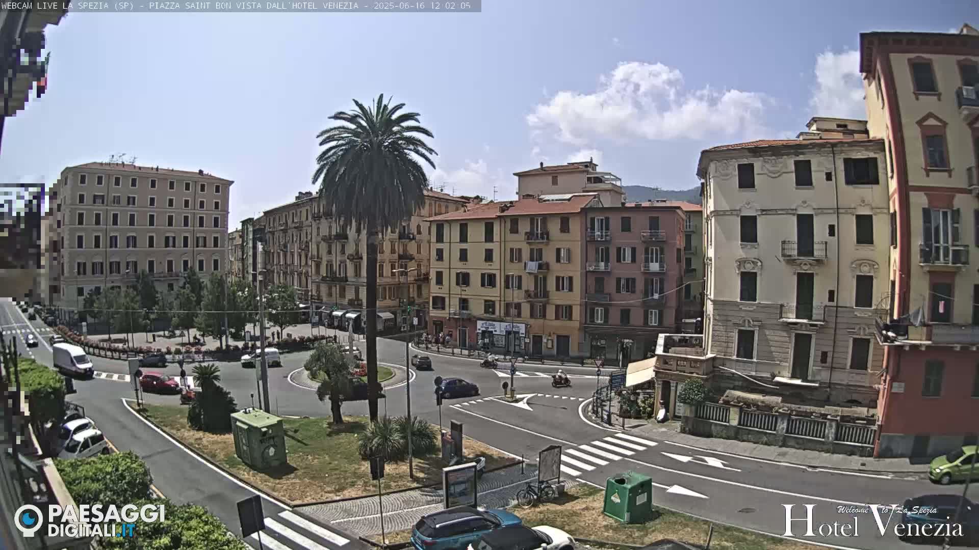 A sunny day reveals a European town square with multi-story buildings surrounding a traffic circle, a tall palm tree in the center, and cars driving on the roads.