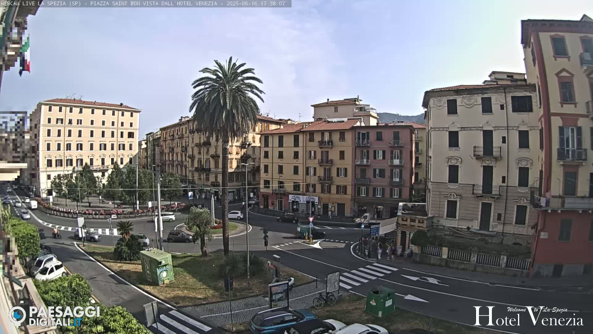 A sunny day in a European city square, showing multi-story buildings surrounding a roundabout with cars and pedestrians.