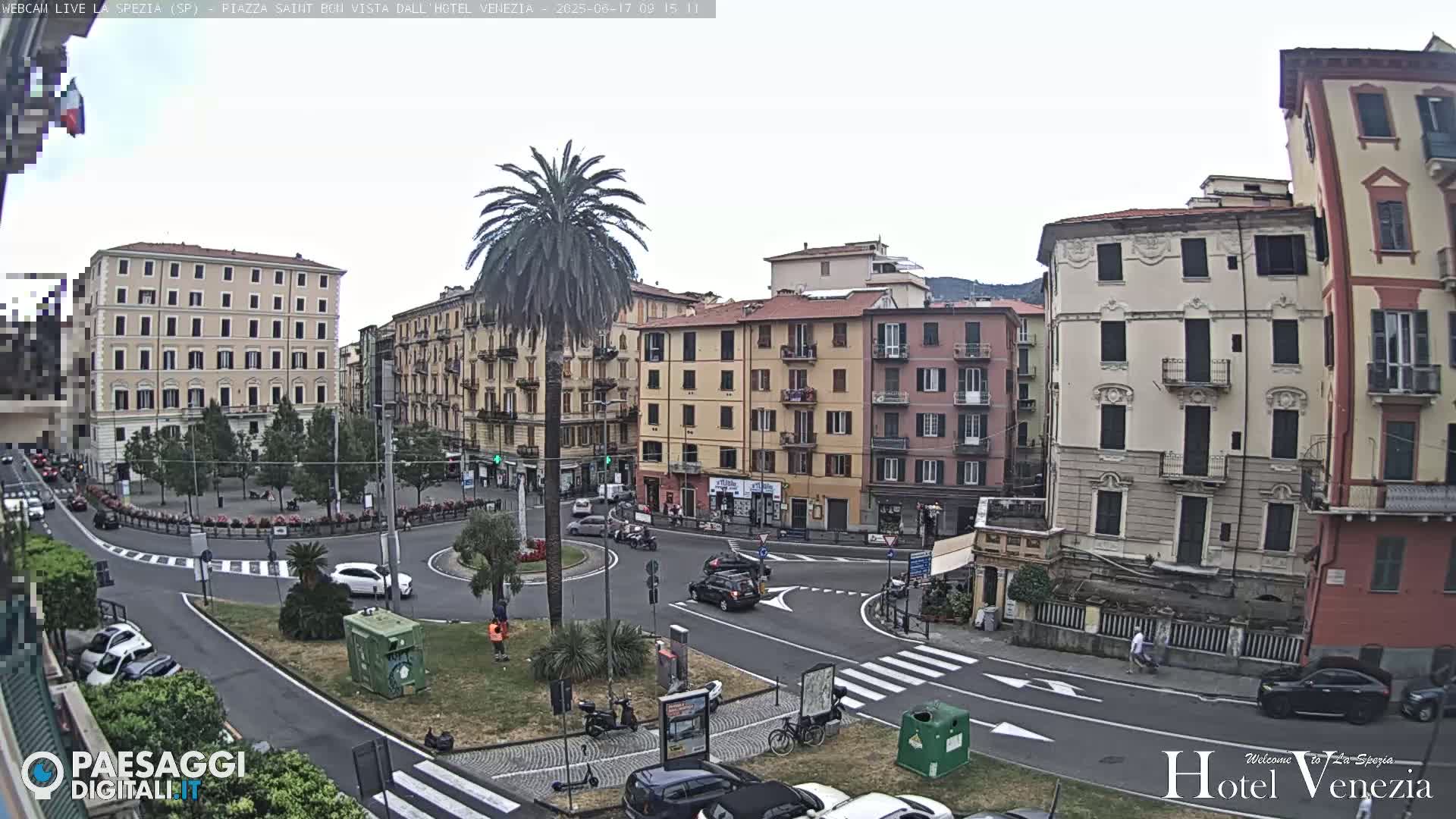 A mostly sunny view of a European town square, with several buildings surrounding a roundabout where cars and scooters are parked or driving.